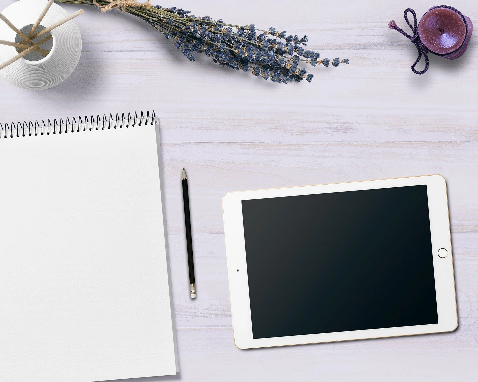 Flat lay of a workspace featuring a tablet with a black screen, a black pencil, a spiral-bound notebook with blank pages, a bunch of dried lavender, a purple candle with a black string, and a white round container with wooden sticks, all arranged on a light wooden surface.