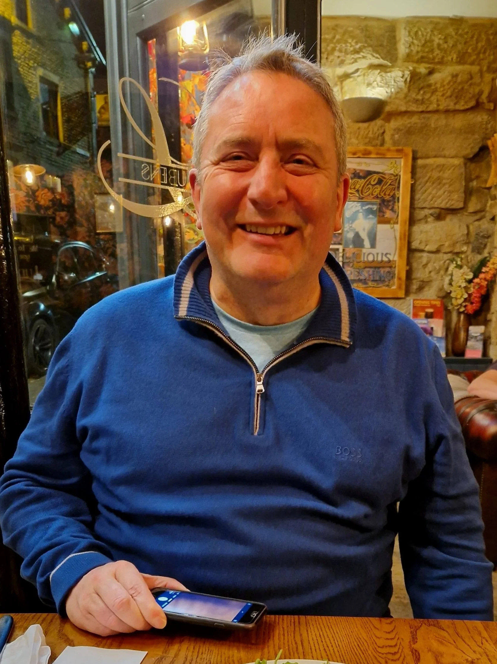 A man with gray hair and a big smile sitting at a wooden table in a cozy indoor setting, wearing a blue quarter-zip sweater, holding a smartphone in his right hand. The background decor includes stone walls, framed pictures, and warm lighting.