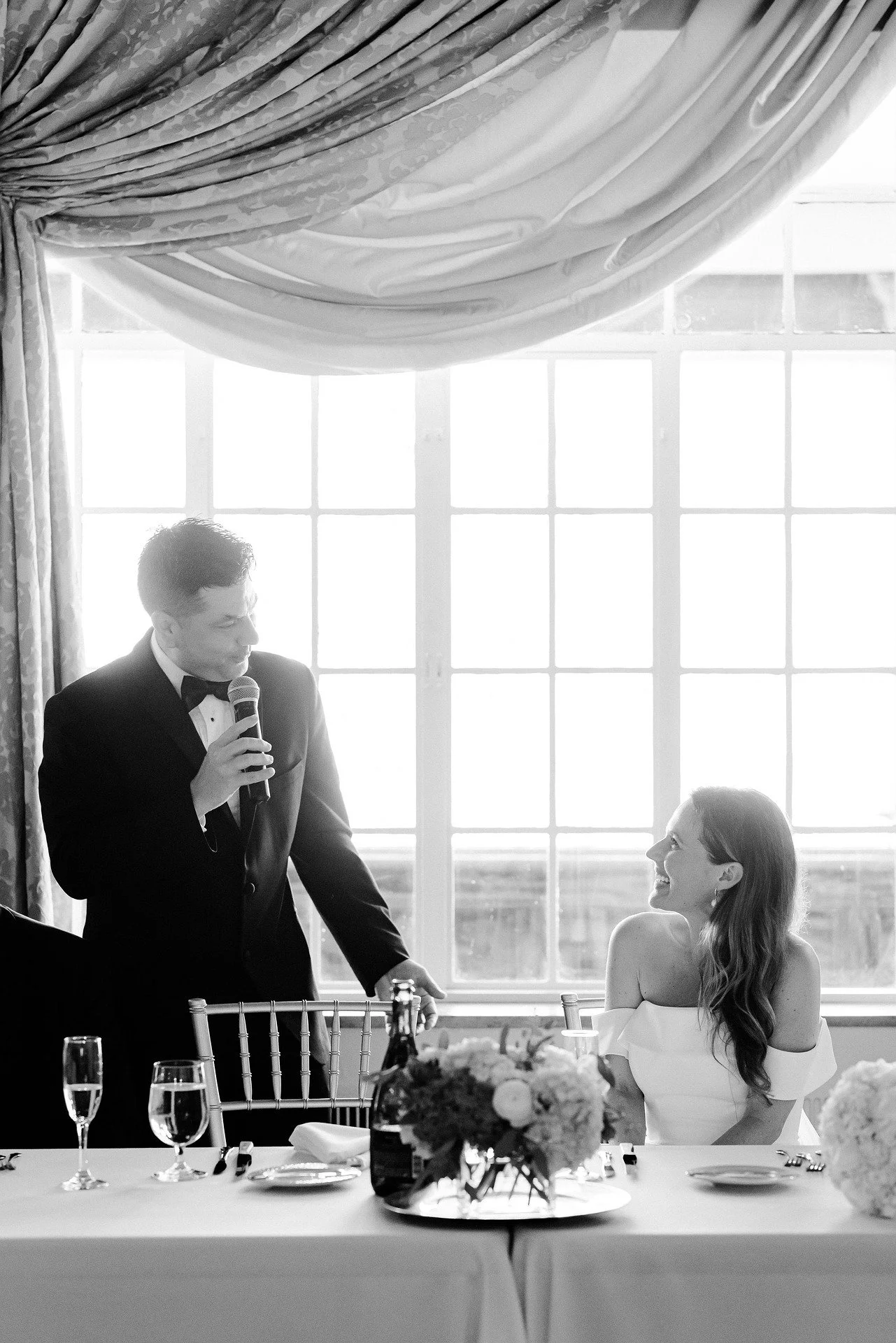 A black and white photo of a wedding reception. A groom in a tuxedo is giving a speech while holding a microphone. The bride, sitting at the table, is smiling and looking at the groom. The table has champagne glasses and a floral centerpiece. Large windows with curtains are in the background, allowing natural light to fill the room.