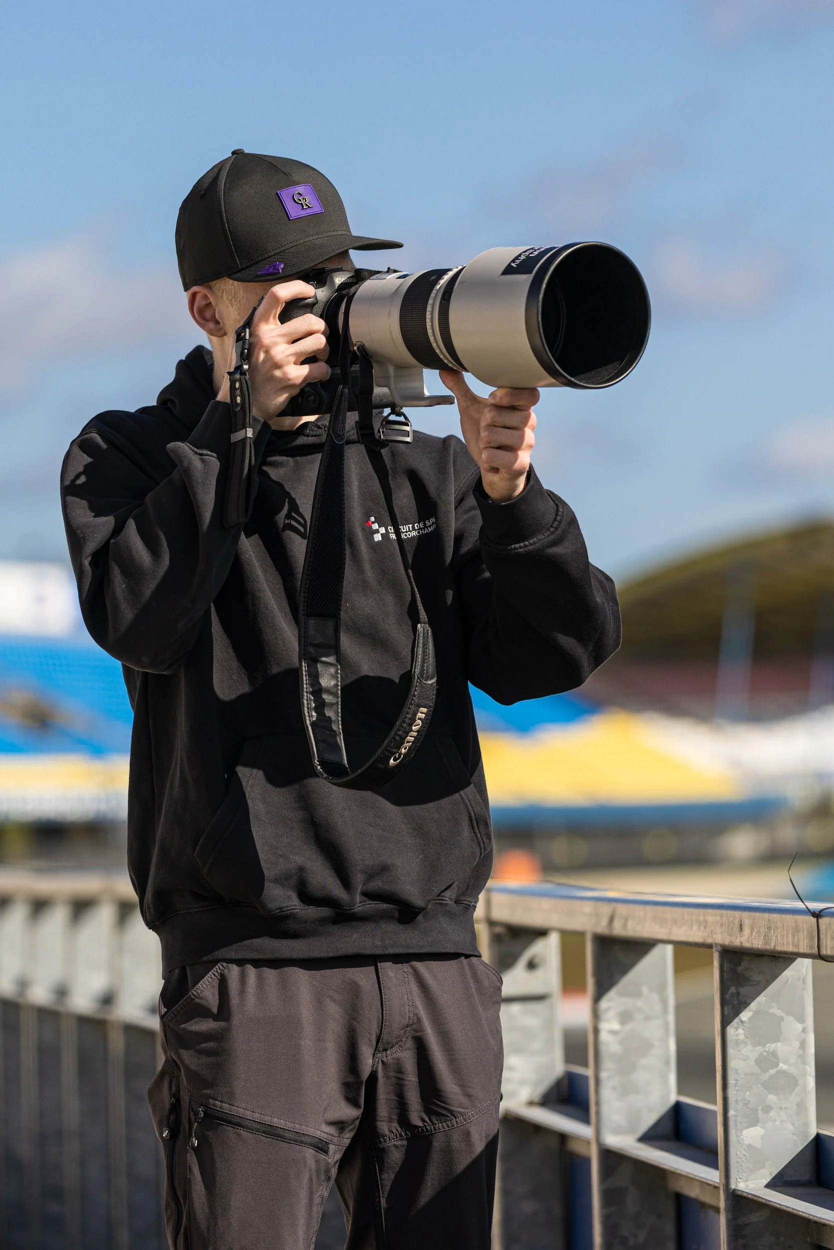 Person wearing black hoodie and dark pants, holding a large camera with a long telephoto lens, taking a photograph outdoors under a blue sky.
