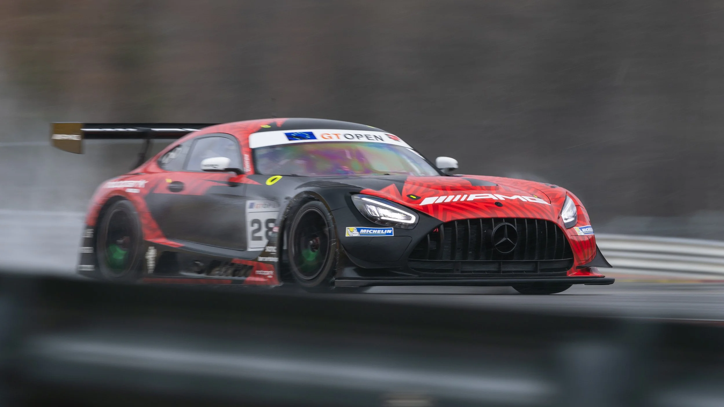 A Mercedes-AMG race car on a wet race track during the GT Open event, with a prominent rear wing and red and black livery.