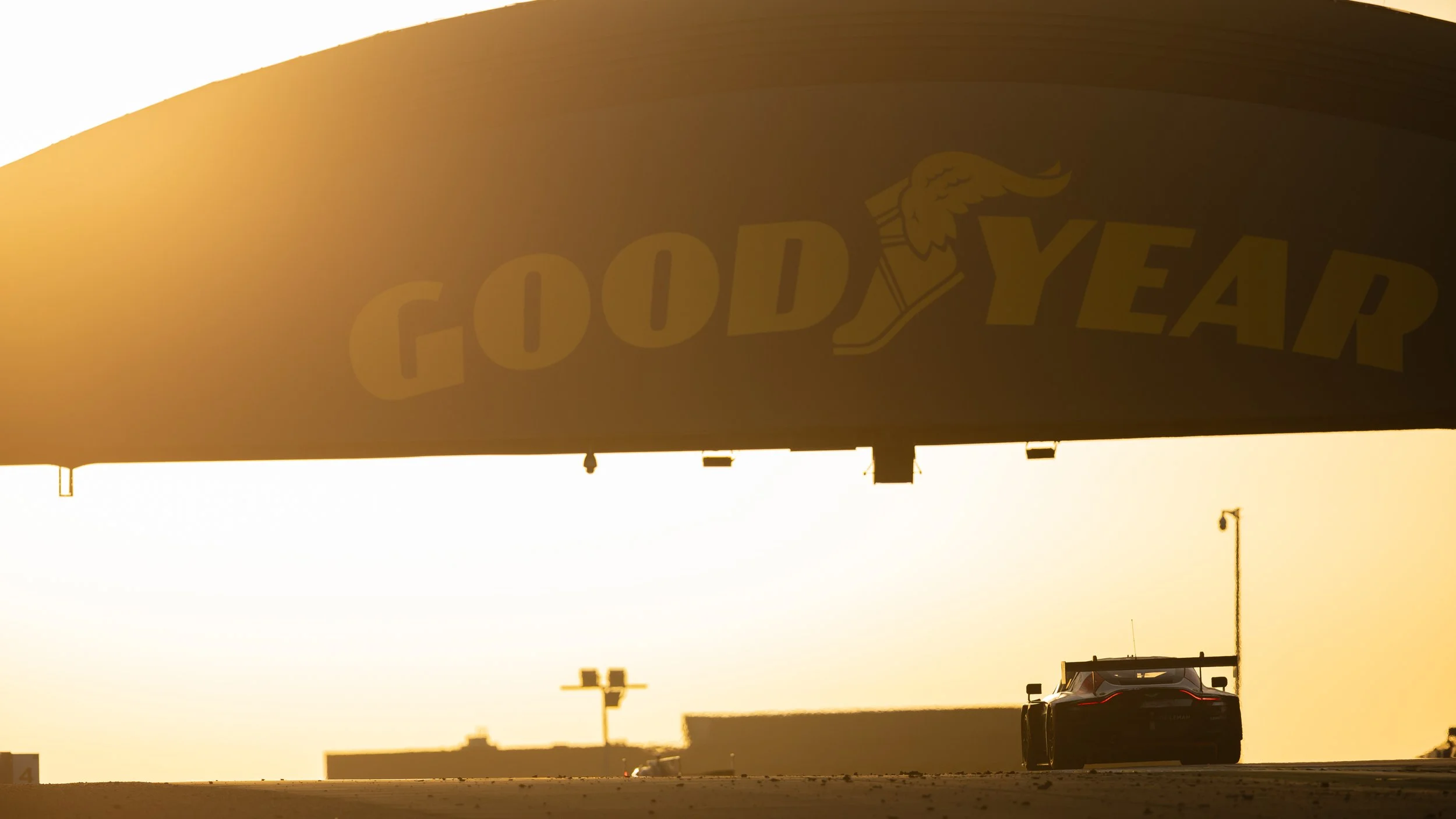 A large billboard with the Goodyear logo and a winged foot symbol, illuminated with sunlight, with a car driving underneath.