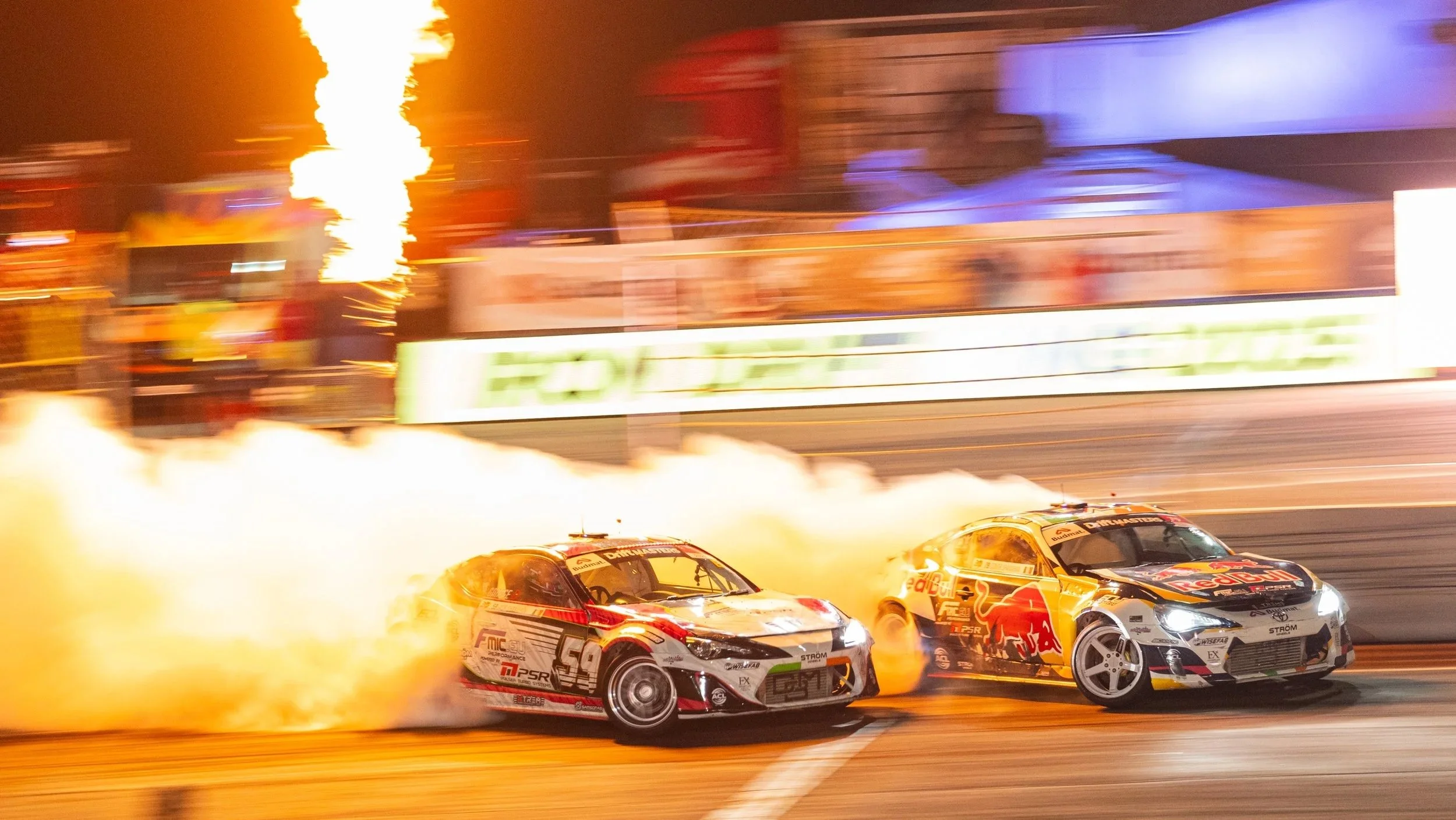 Two race cars drifting on a track at night, with flames and sparks coming from the tires, and a background with blurred signage and bright lights.