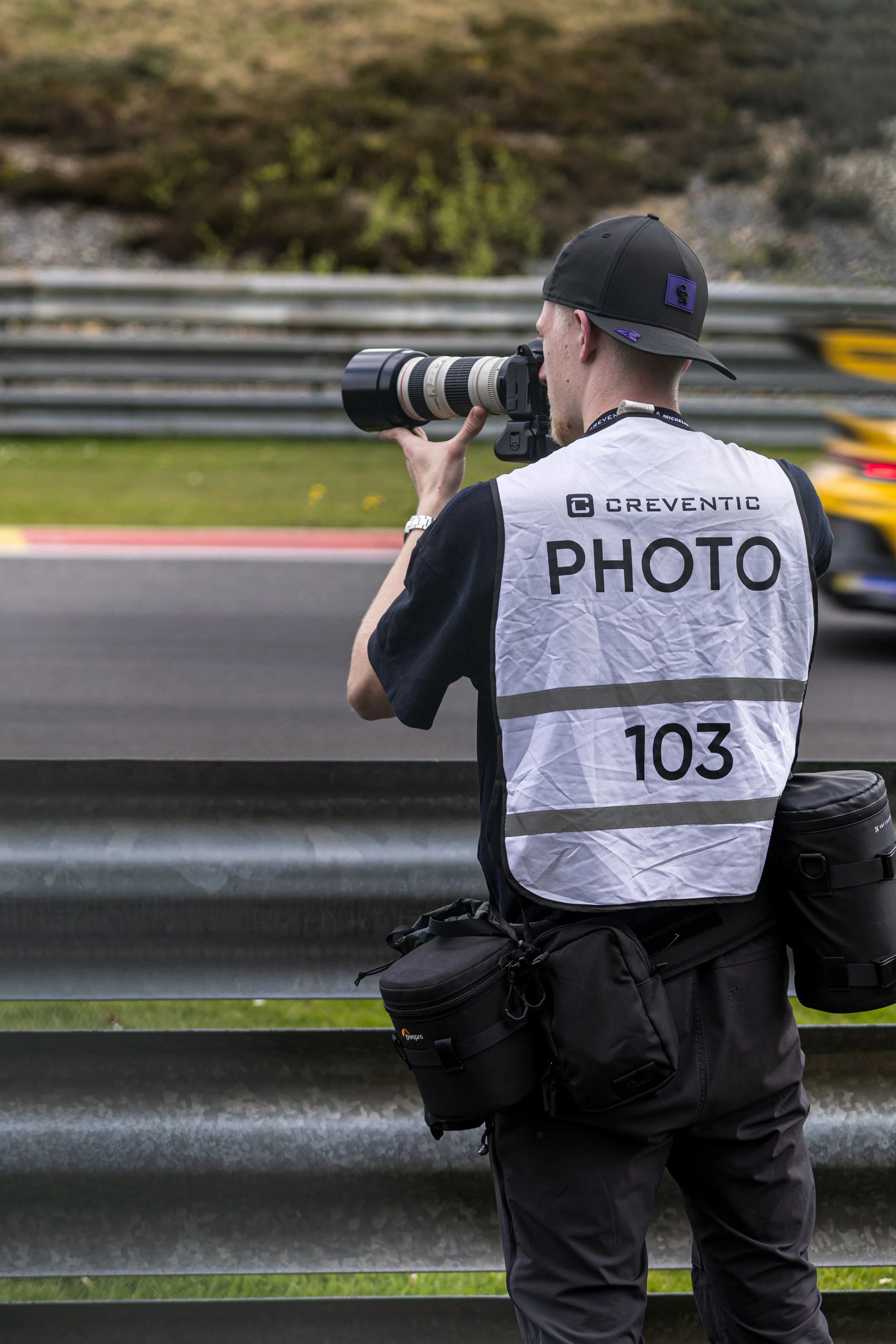 A photographer wearing a white vest with 'PHOTO' printed on it, holding a camera with a large telephoto lens, and kneeling near a guardrail on a race track.