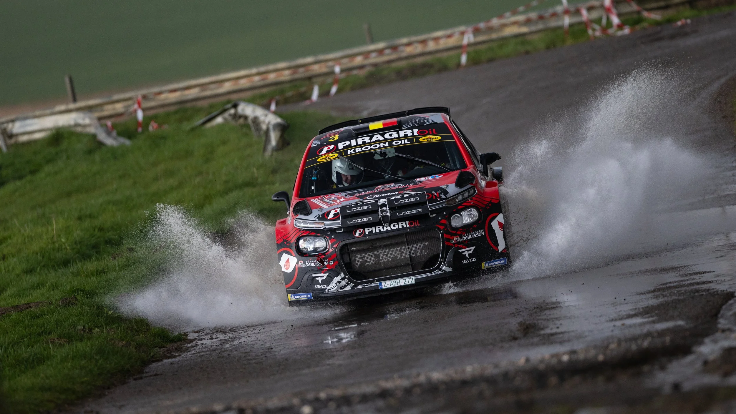 A rally car speeding on a wet dirt road, creating a spray of water and mud, with grass on the side and a guardrail in the background.