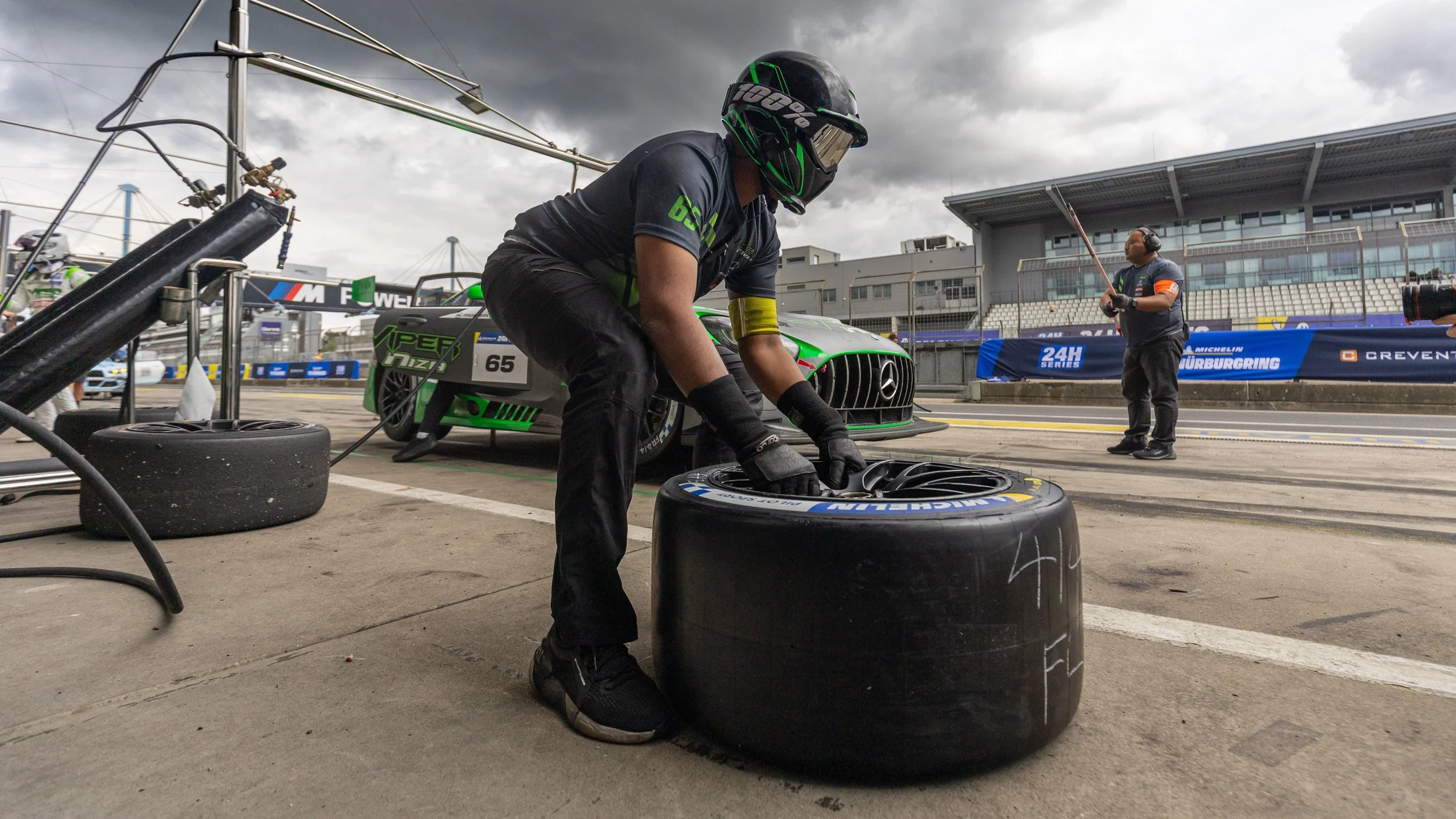 Race car crew member changing a tire in the pit lane during a race, with a green race car and crew member in the background under a cloudy sky.