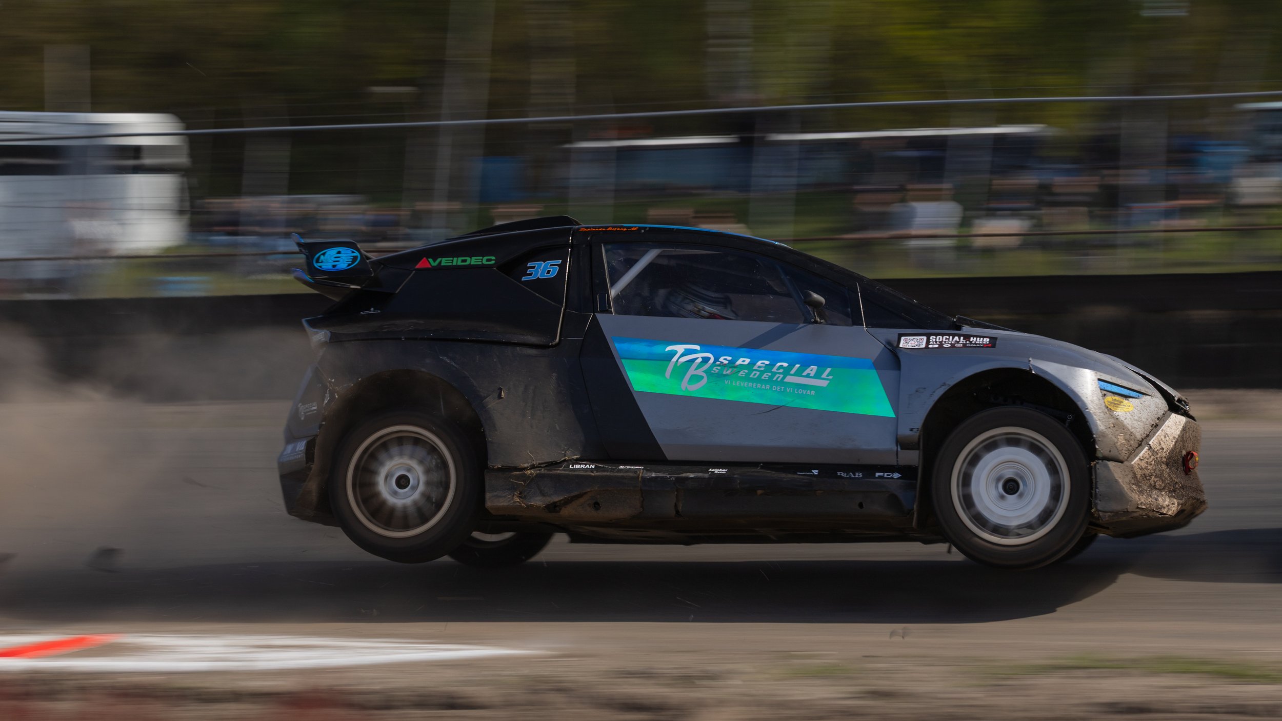 A black race car with various sponsorship logos speeds along a dirt track, kicking up dust behind it.