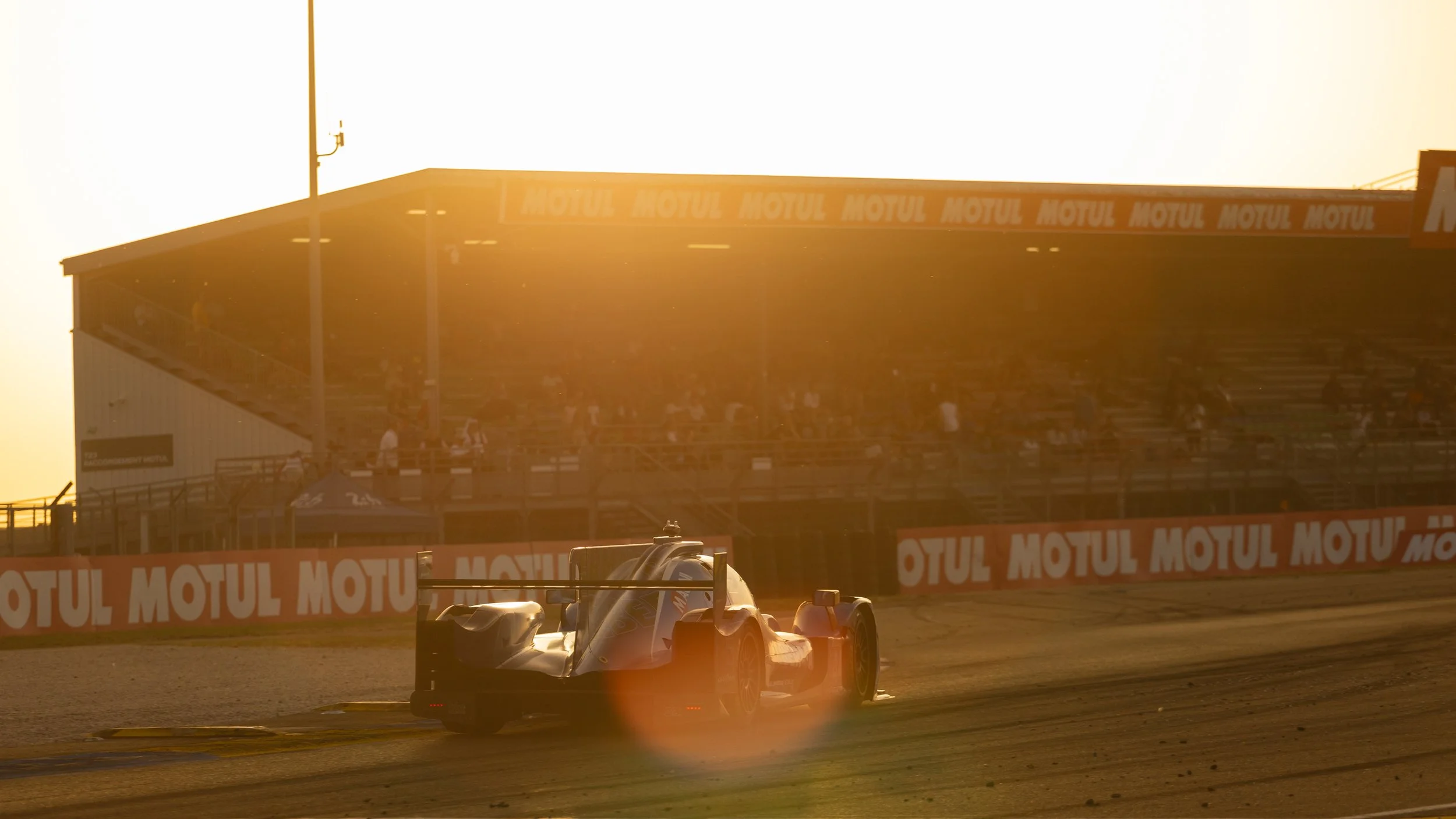 Race car driving on a track at sunset with grandstands and Motul sponsorship banners in the background.