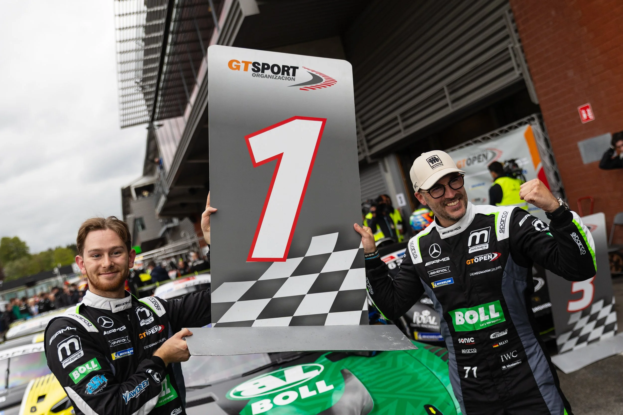 Race car drivers celebrating on the podium after a race, holding a large checkered flag and a sign that says '1', with other race cars and spectators in the background.
