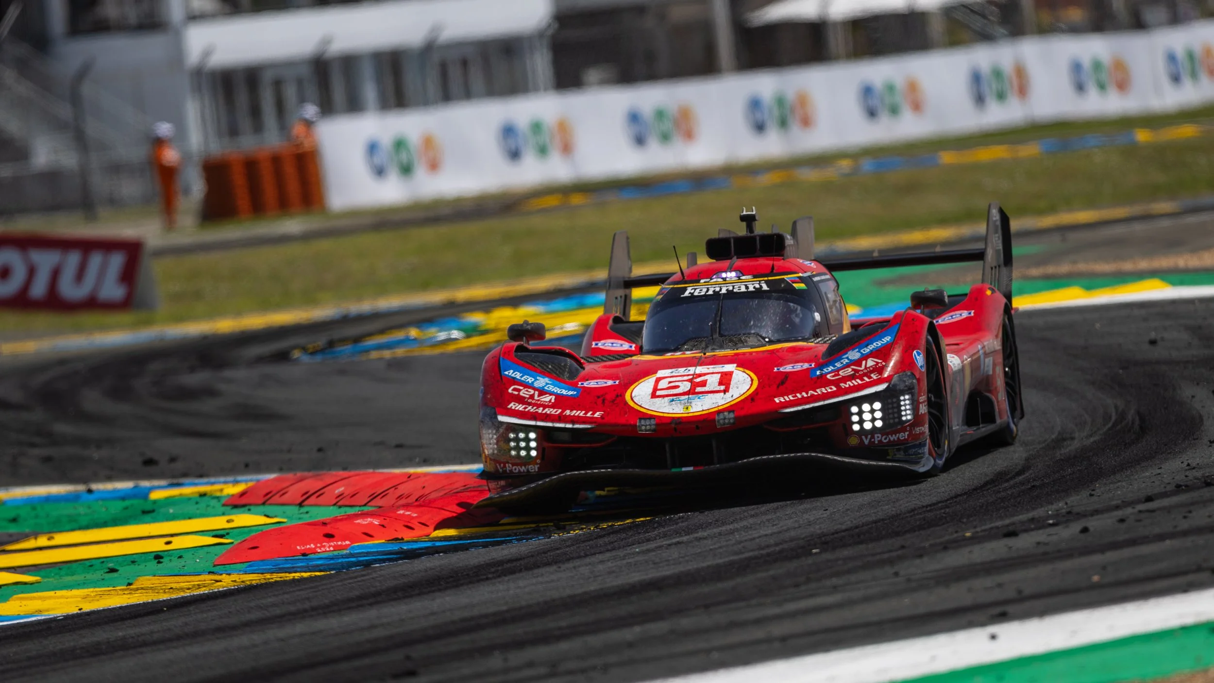 A red race car on a racetrack, with a blurred background of safety barriers and trackside advertisements.