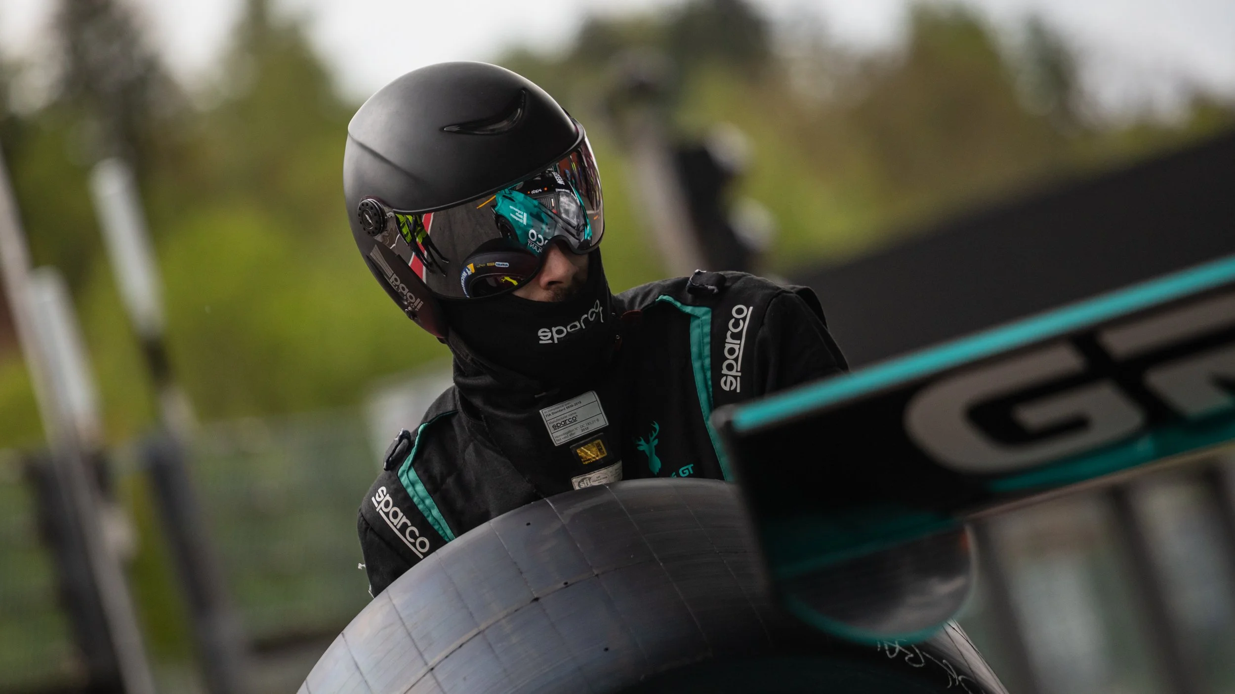 A race car driver wearing a black helmet with a reflective visor, a black racing suit with teal accents and sponsors, is preparing for a race near a tire barrier.