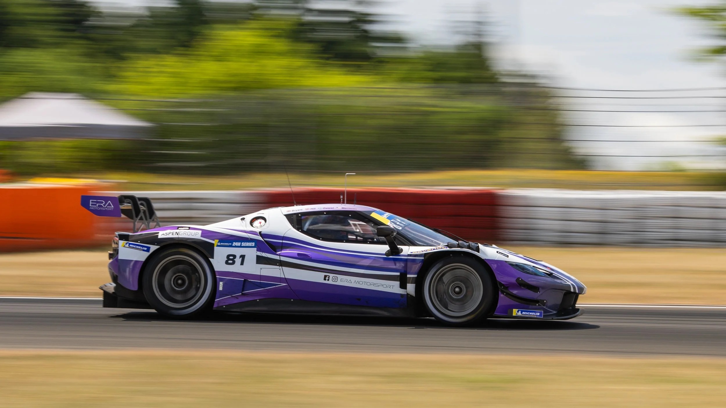 A purple and white race car with the number 81 speeds along a race track with blurred background trees and barriers.