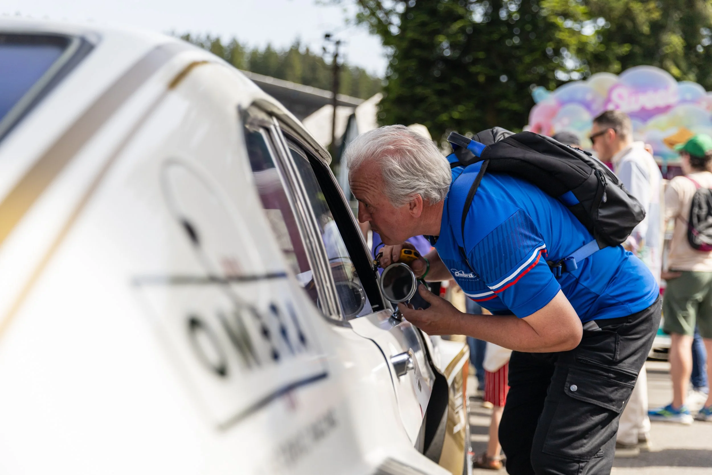 An older man with white hair, wearing a blue shirt and a backpack, reading a document or ticket through the window of a white car at an outdoor event, with people and colorful balloons in the background.