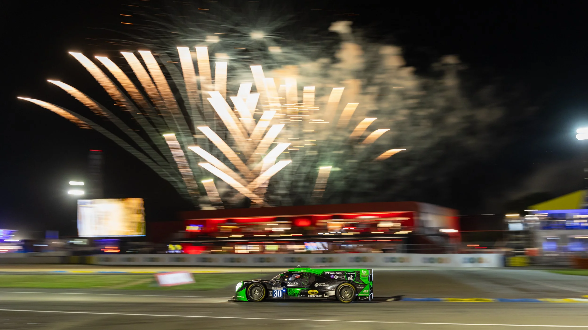 Nighttime scene at a race track with fireworks in the sky, a race car with the number 30 on it on the track, and blurred lights and structures in the background.
