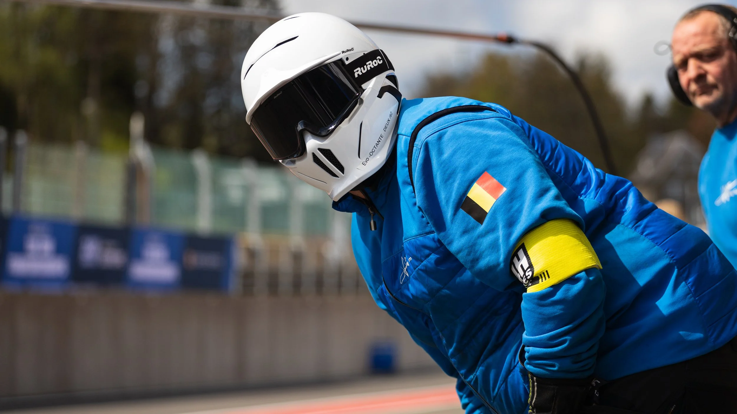 A mechanic car driver wearing a blue racing suit with a Belgian flag patch on the sleeve, a yellow armband, and a white helmet with black visor, leaning forward at a racing event, with a man in a blue shirt and headphones in the background.