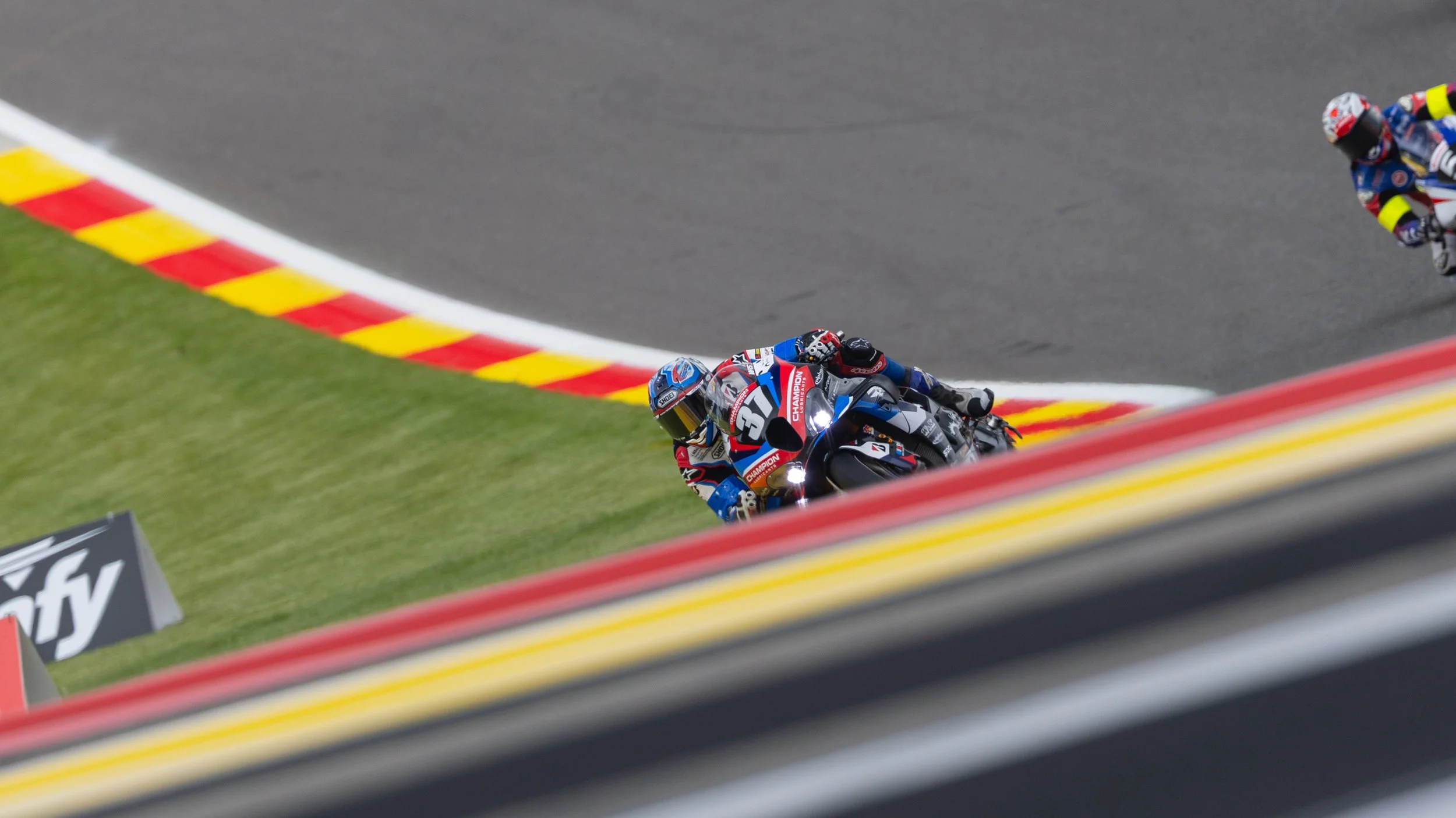Motorcycle racer leaning sharply into a turn on a race track, with a second rider visible in the background. The track features red and yellow curb markings.