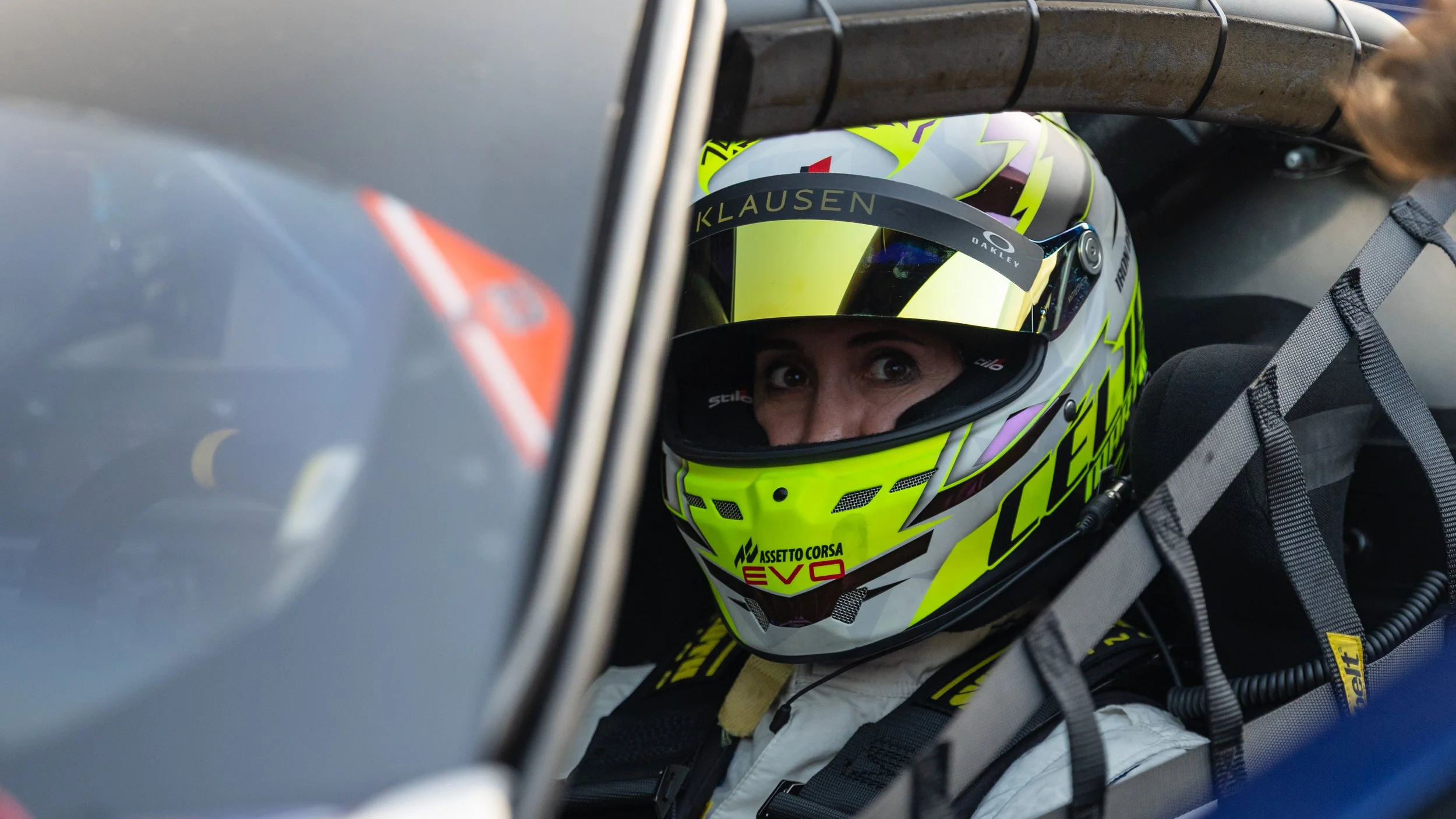 Close-up of a race car driver wearing a bright yellow helmet with black and purple details, looking through the visor from inside the car, with safety gear and equipment visible around.