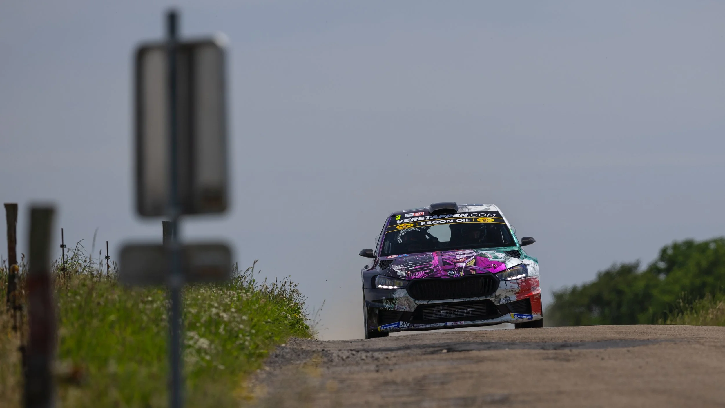 A race car with colorful pink, purple, and black graphics speeds down a gravel track, kicking up dust against a overcast sky.