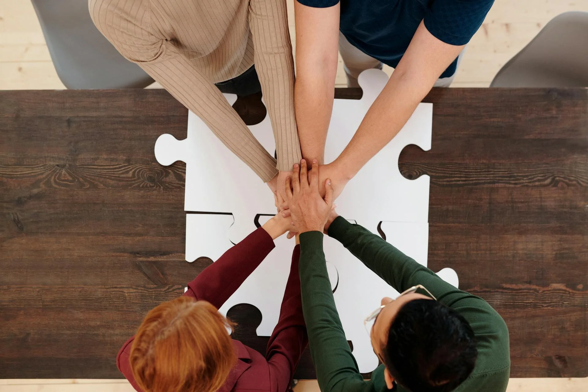 Un groupe de cinq personnes se tient autour d'une table en bois, joints par leurs mains posées sur un grand puzzle blanc formant une pièce, symbolisant la collaboration d'équipe.