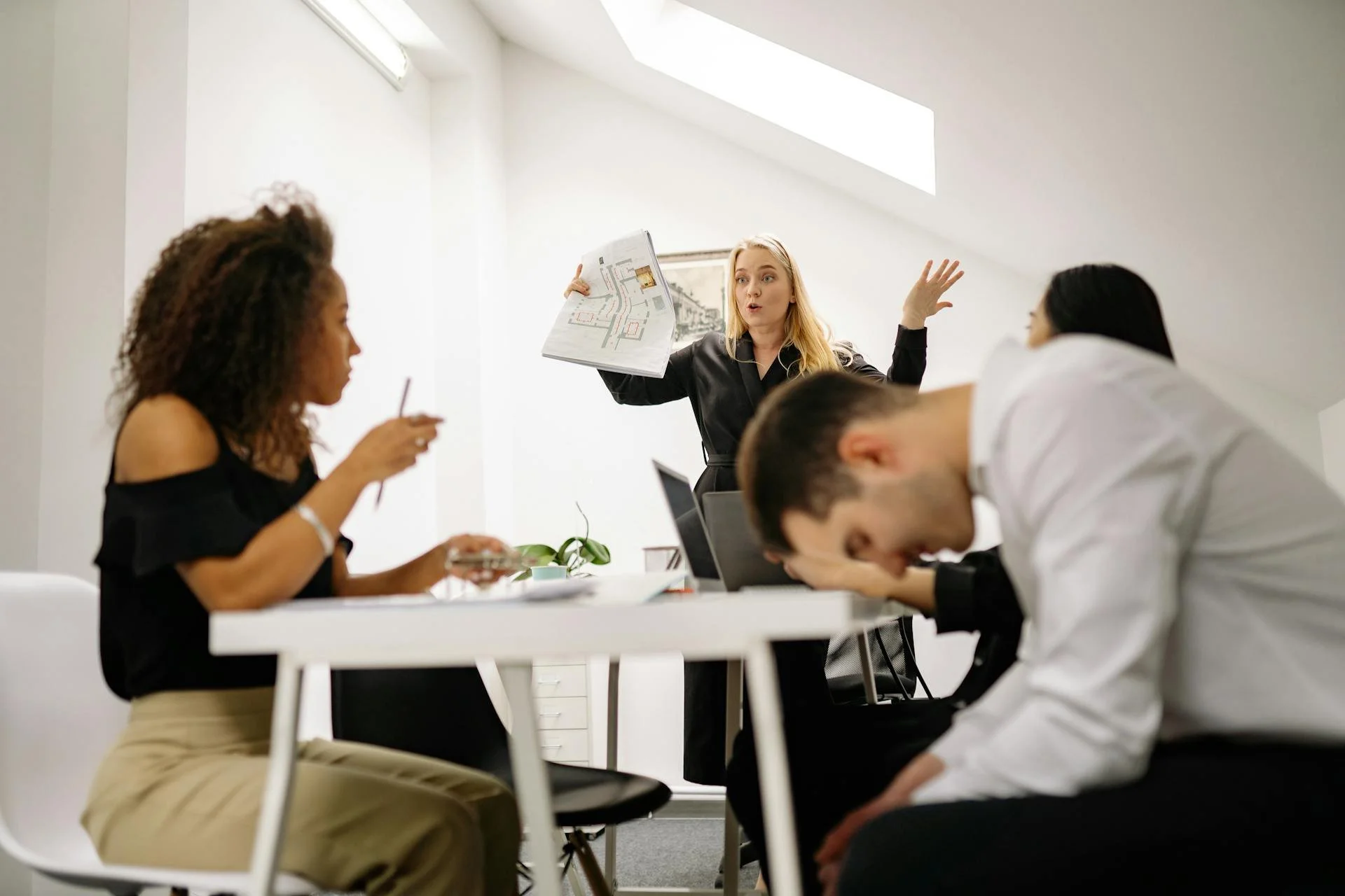 Une femme blonde en blouse noire tient un plan ou une carte et parle à un groupe de quatre personnes assises, dont deux hommes. Une femme à gauche, en haut, utilise un téléphone, et un homme à droite, en bas, a la tête inclinée sur la table, apparemment fatigué ou pensif. La scène se déroule dans une pièce lumineuse avec un plafond en pente et un éclairage naturel.