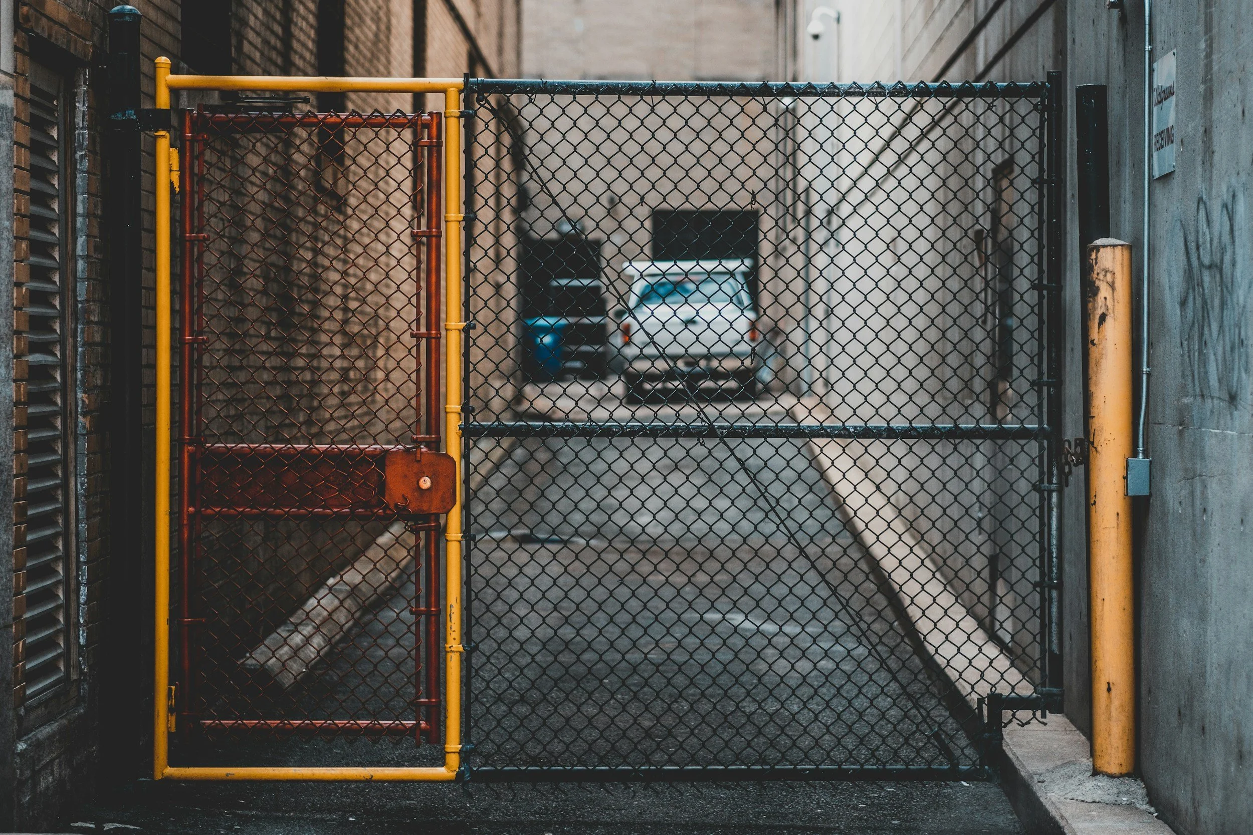 A metal gate with a chain-link fence blocking the alleyway, with parked cars visible in the background.