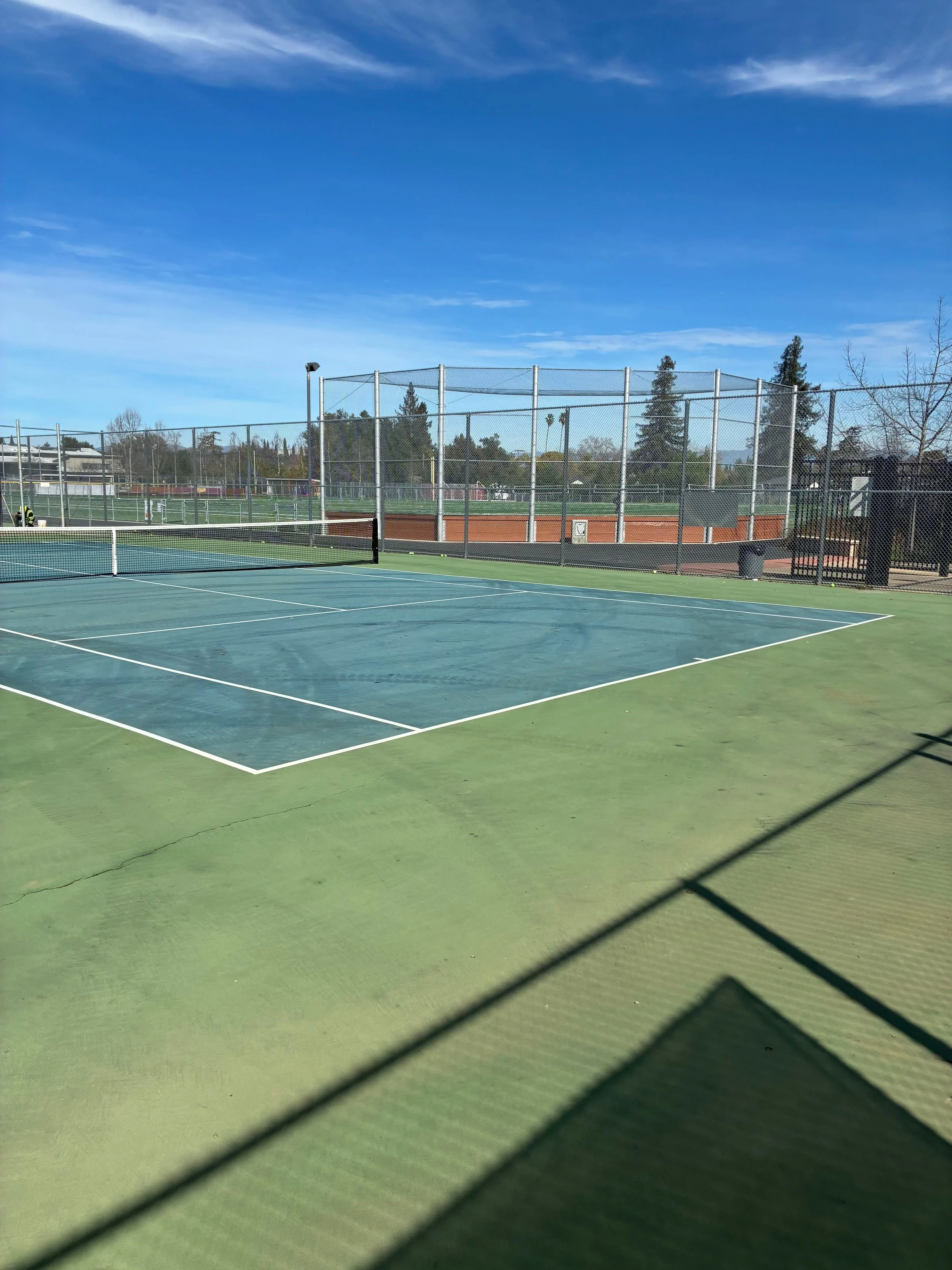 Empty tennis court under a blue sky with some clouds, surrounded by chain-link fences and trees in the background.