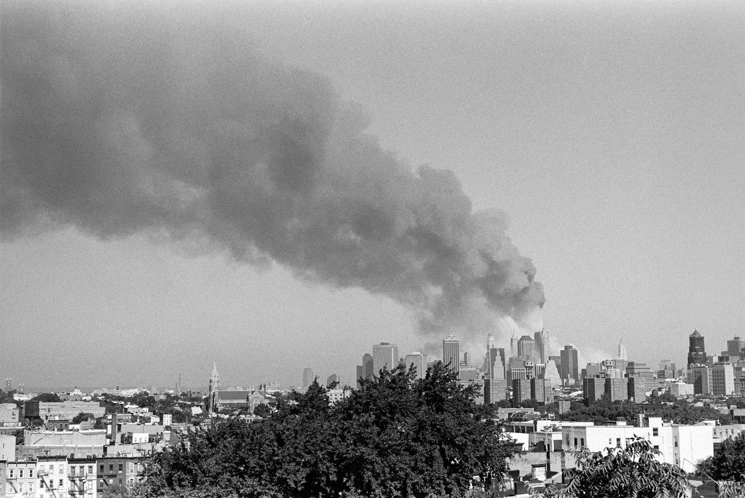 Black and white photo of a city skyline with smoke billowing from a building amid tall skyscrapers and lower buildings in the foreground.