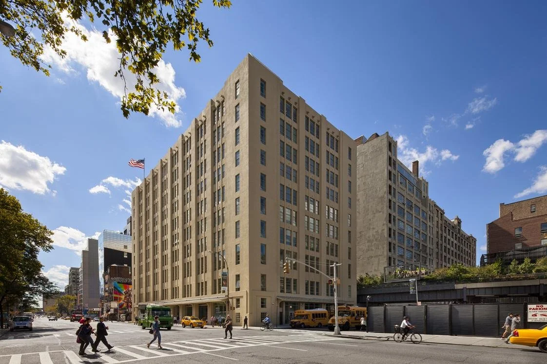 City street scene with pedestrians crossing, yellow cabs, a bus, and a large beige building with many windows under a partly cloudy sky.