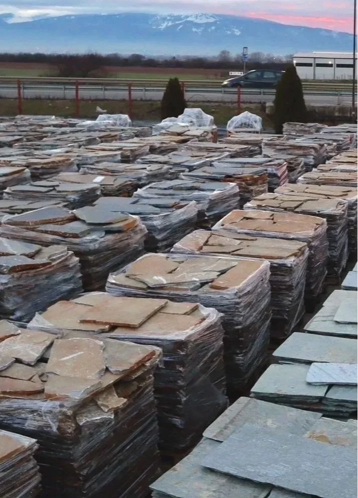 Stacks of cut stone slabs wrapped in plastic outdoors at sunset with a mountain in the background.