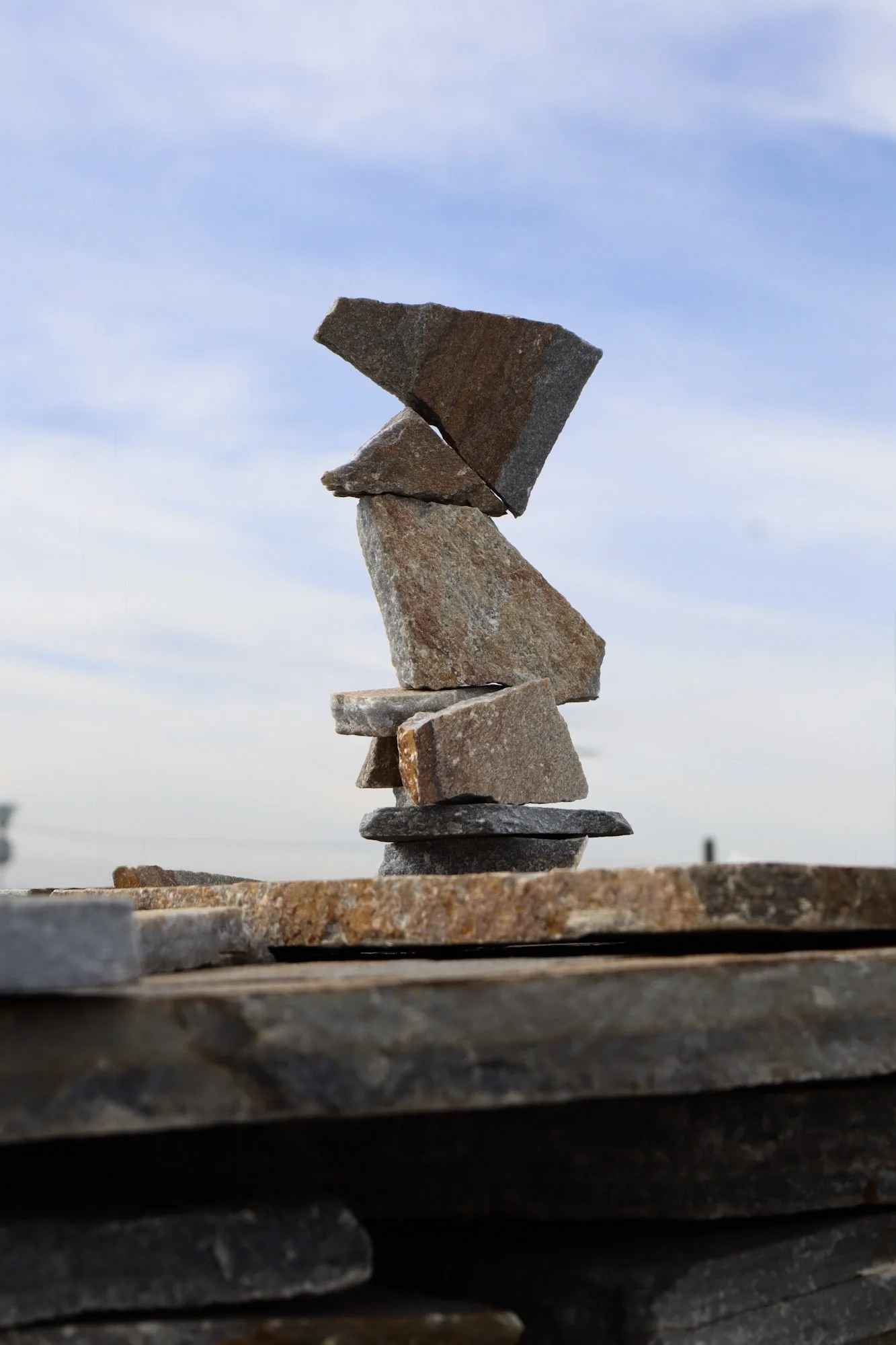 Stacked flat stones forming a balanced tower against a blue sky with scattered clouds.
