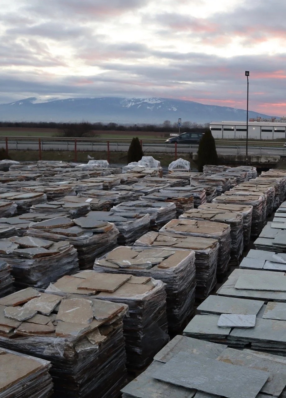 Stacks of flat stone tiles wrapped in plastic outside with mountains and a cloudy sky in the background.