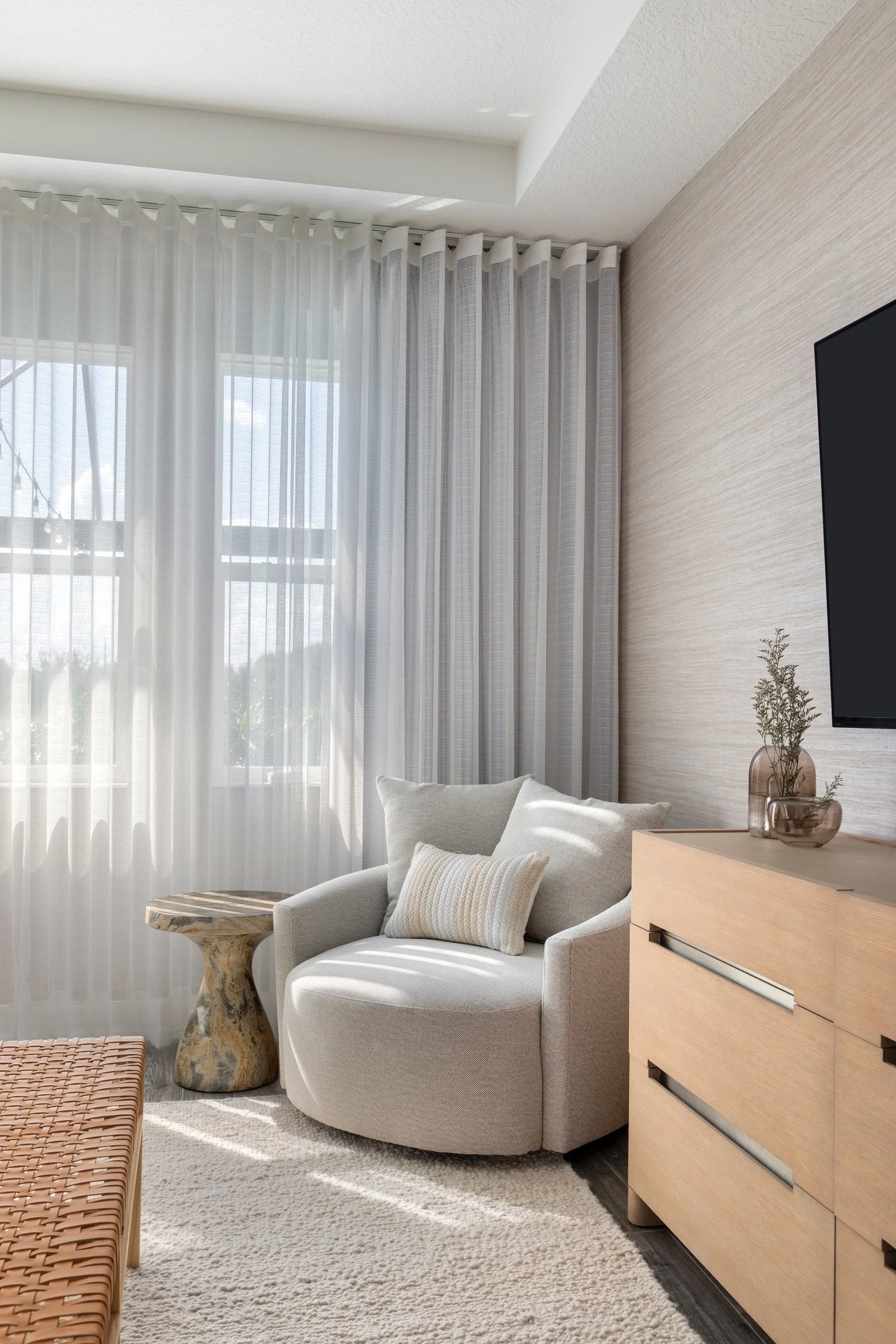 Bright living room with sheer curtains, a white armchair with a pillow, a wooden side table, a flat-screen TV, and decorative plants.