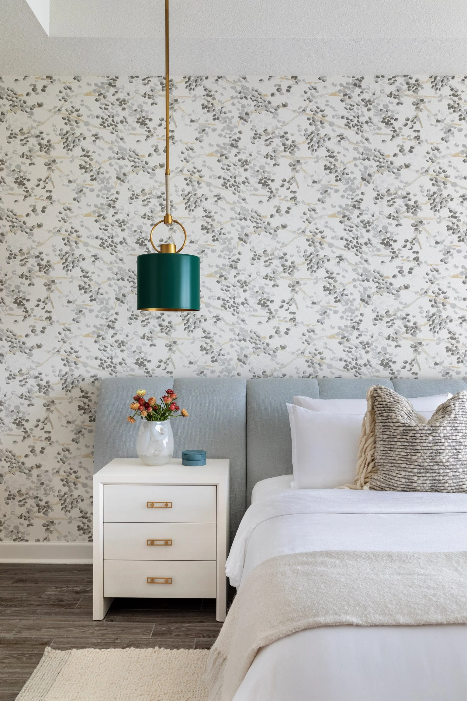 A bedroom with a white nightstand, a vase of flowers, a small blue container, and a bed with pillows. The wall has patterned wallpaper, and a green pendant light hanging from the ceiling.