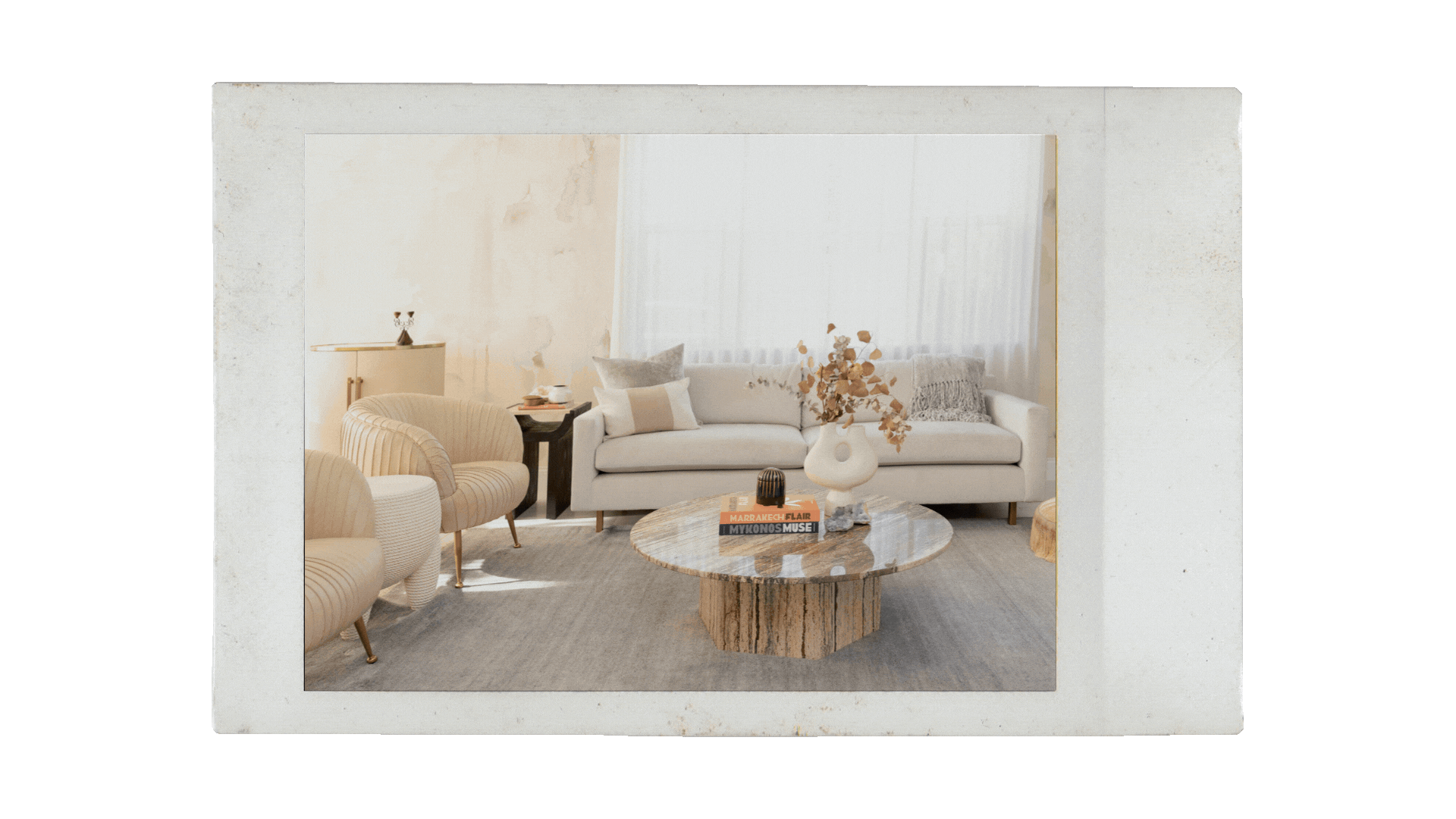 A modern living room with white sofas, a beige armchair, a round marble coffee table, and decorative items including books, a white vase with dried flowers, and a black sculpture, all against a white wall with sheer curtains.