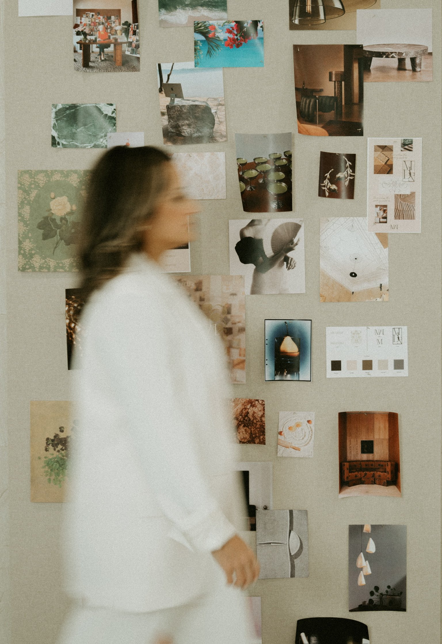 A woman in a white blazer walking past a wall with various photographs and art prints pinned to it.