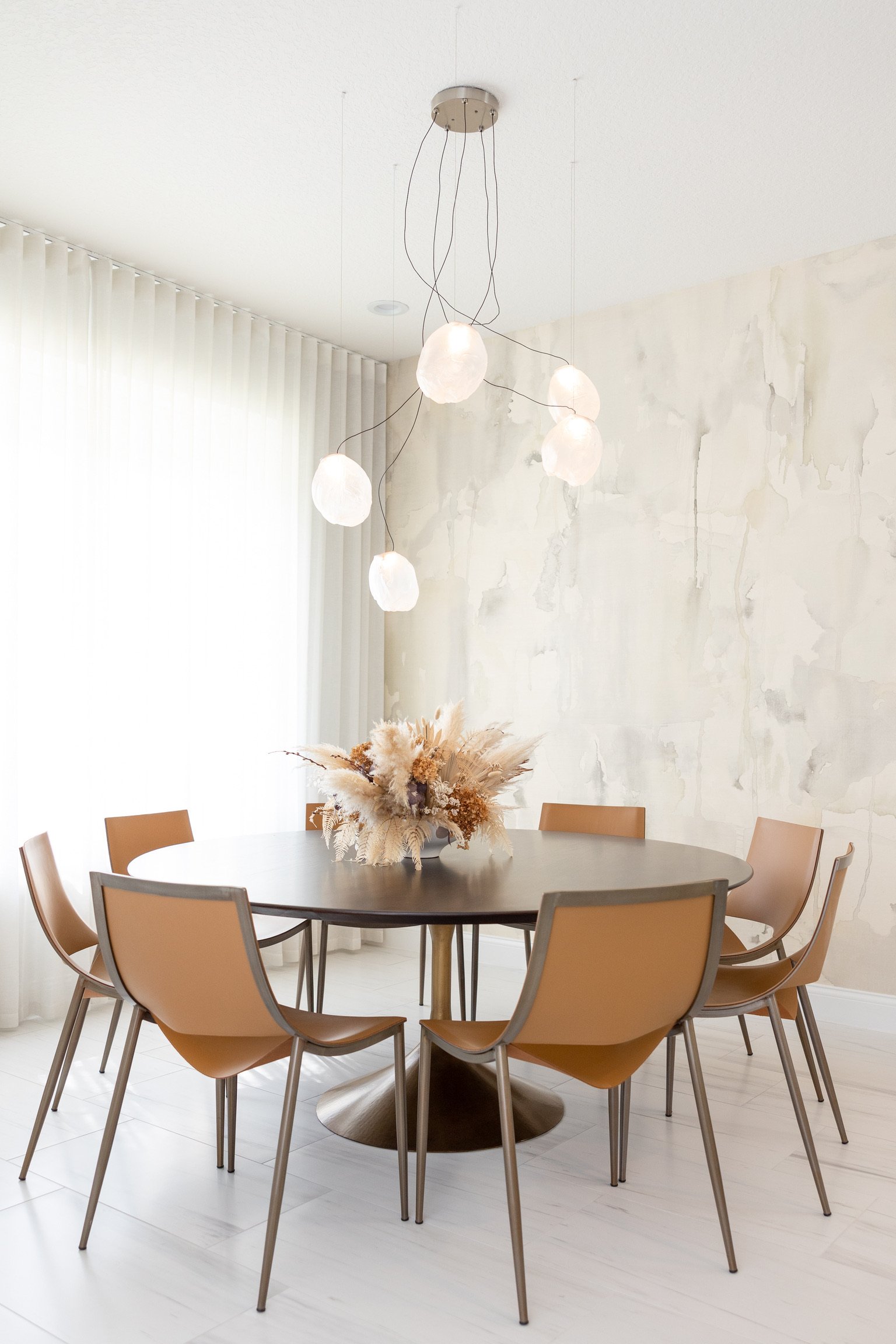 Round dining table with six tan chairs, a large bouquet of beige dried flowers as a centerpiece, and a modern chandelier with six white spherical lights hanging from the ceiling in a bright dining room.