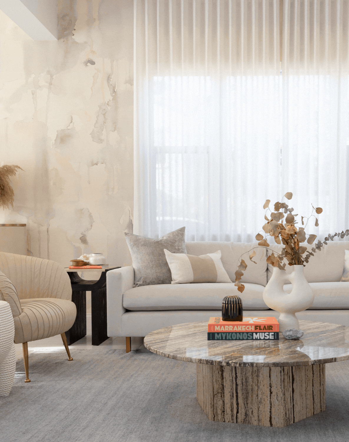 A cozy living room with neutral-colored furniture, a marble coffee table with books and decorative objects, a white sofa with pillows, armchair, sheer curtains, and a textured wall.