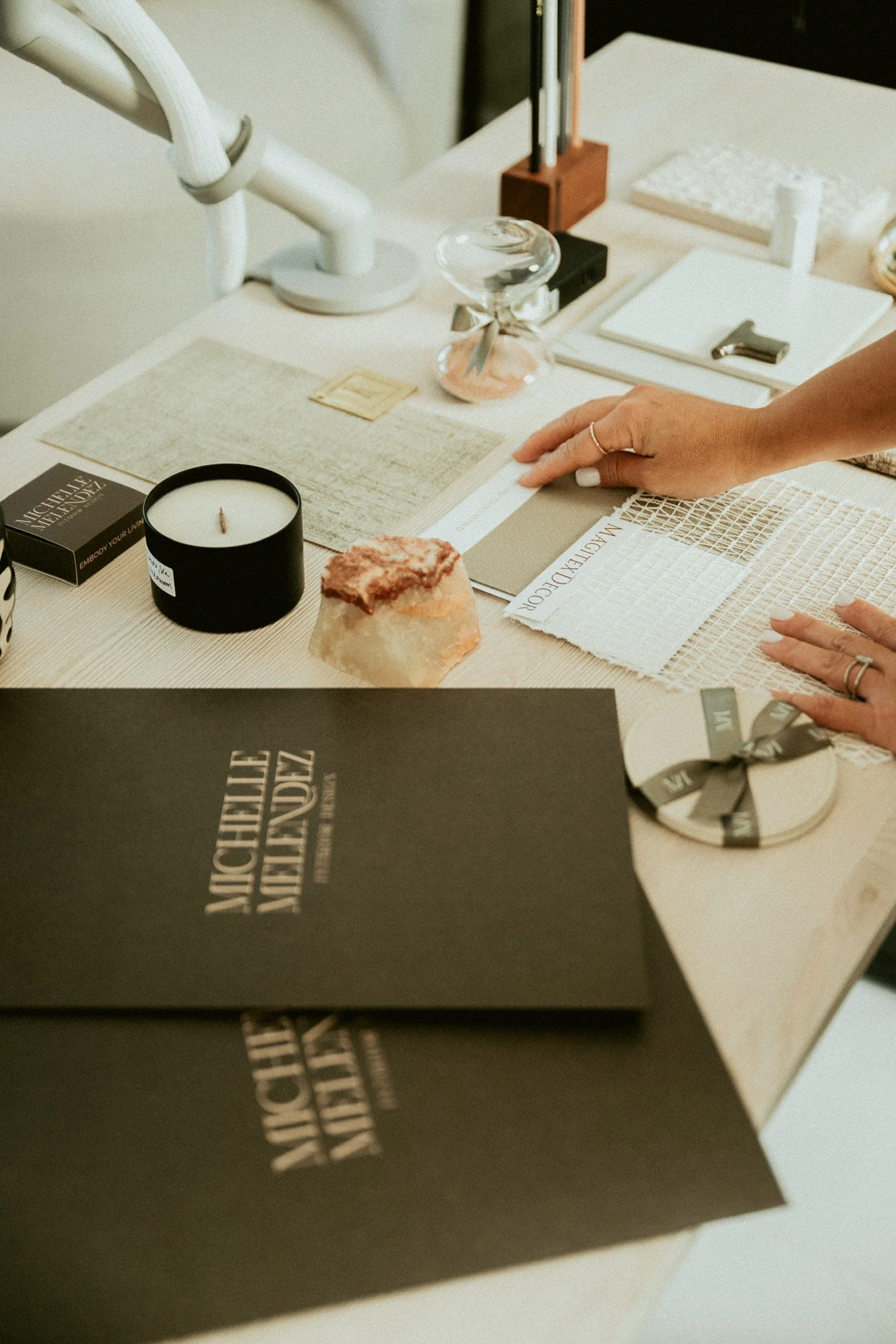 A collection of design and decor samples on a light-colored table, including fabric swatches, a candle, a mineral specimen, and various stationery and packaging items, with someone's hand reaching over.