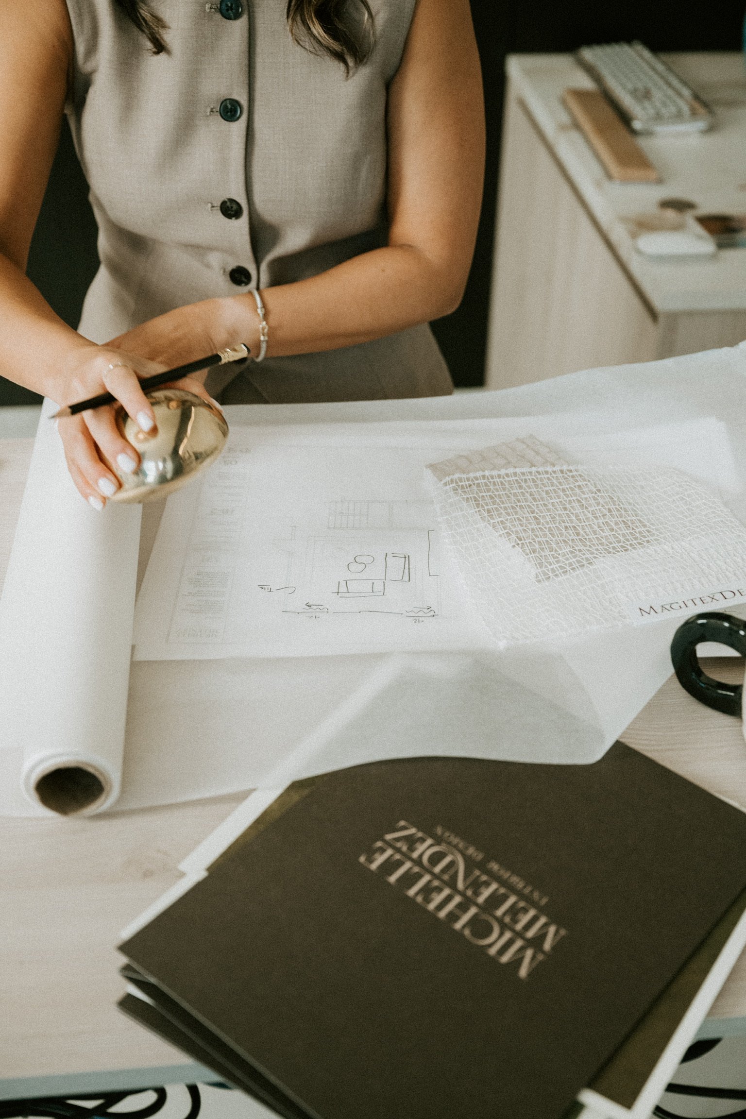 A woman in a beige vest reviewing architectural plans and design samples on a desk, with a black hardcover book titled "ARCHITECTURAL
