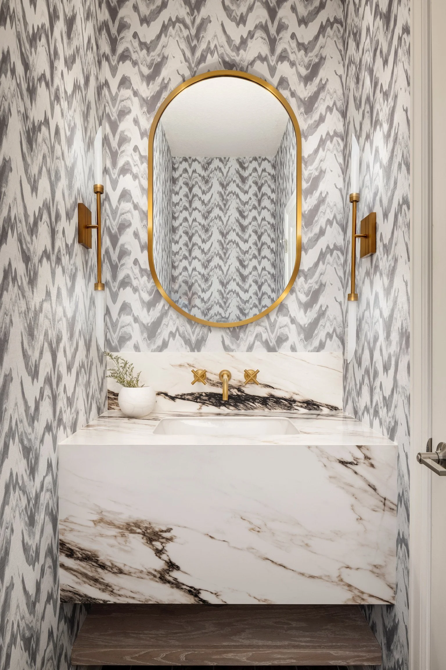 A modern bathroom with a marble sink and a large mirror. The walls are decorated with a zigzag gray and white pattern. Gold fixtures include the mirror frame, sconces, and faucet, with a white potted plant on the sink.