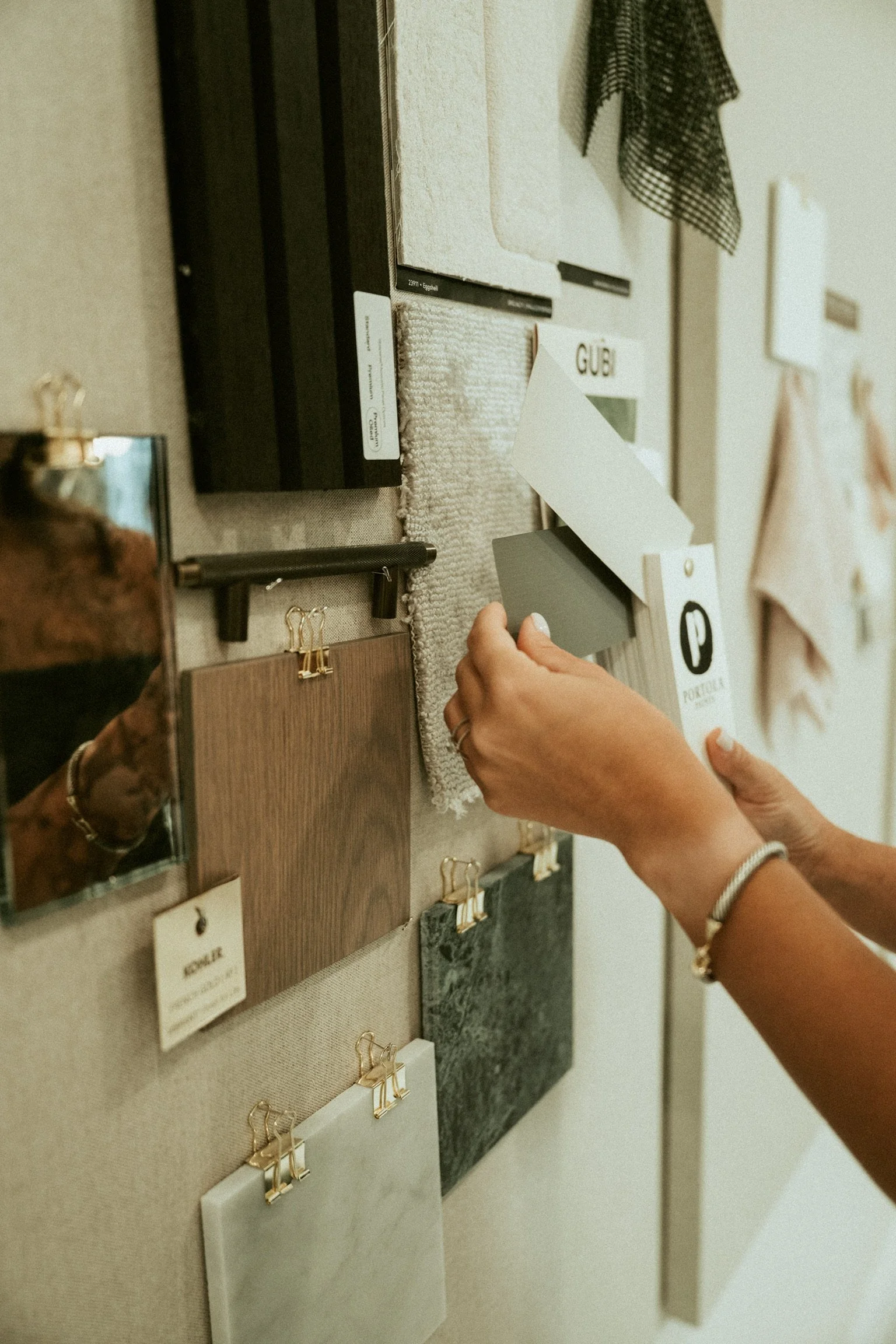Person holding a color sample card in front of a display of various textured material samples on a wall, with some small fabric and paper samples pinned nearby.