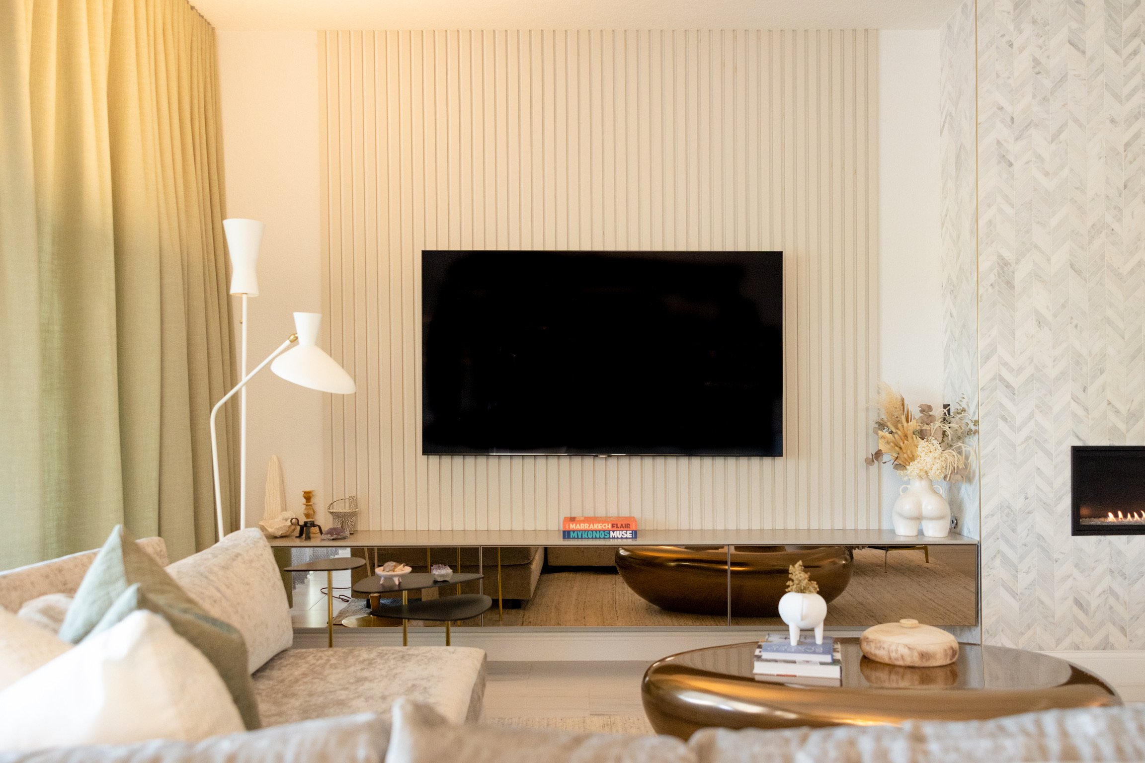 Living room with a wall-mounted flat-screen TV, beige sofa, side table, and modern decor including vases and books.