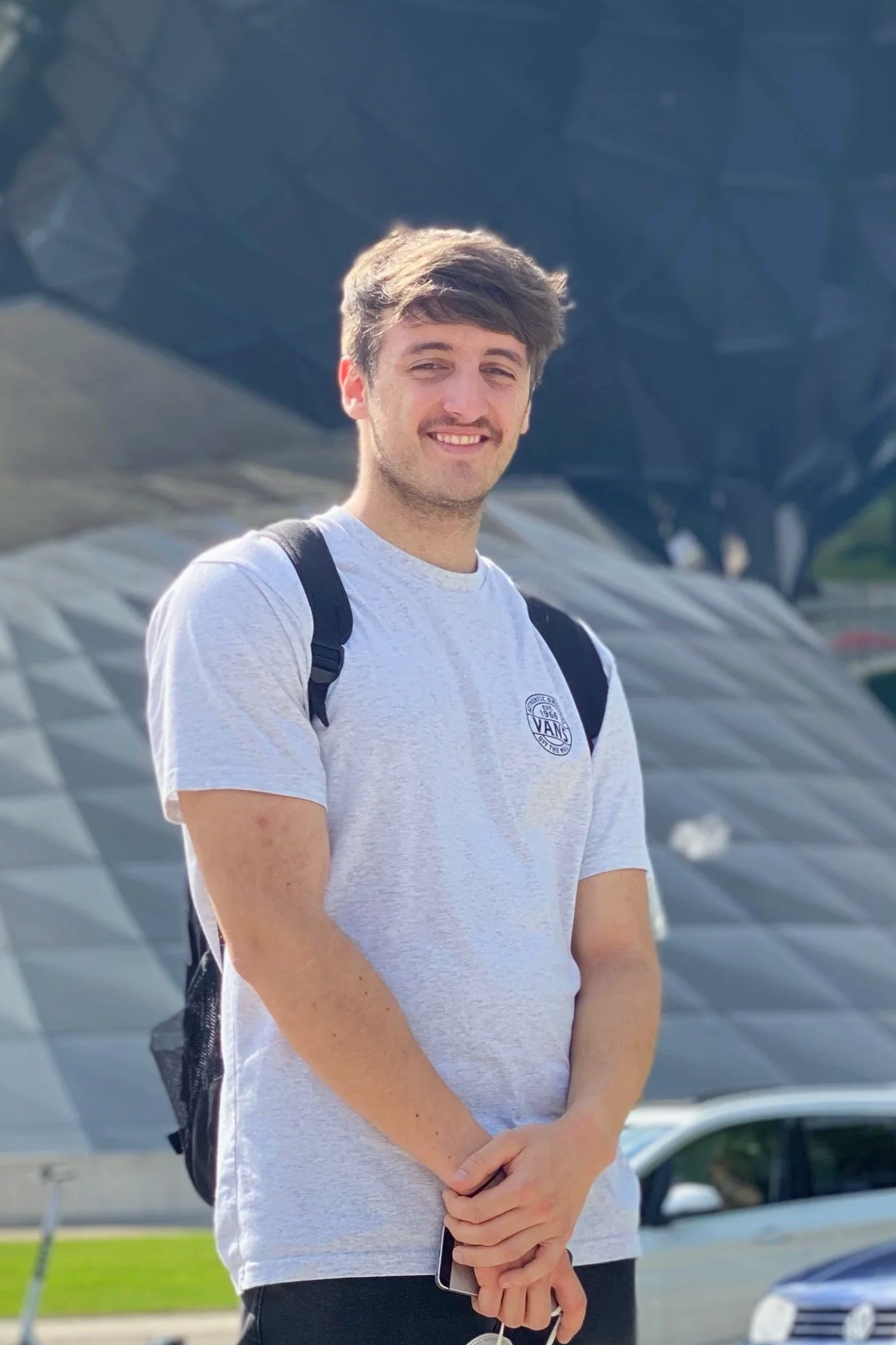 Junge Mann mit grauem T-Shirt und Rucksack steht vor einer modernen Architektur im Freien, sonniger Tag, lächelt.