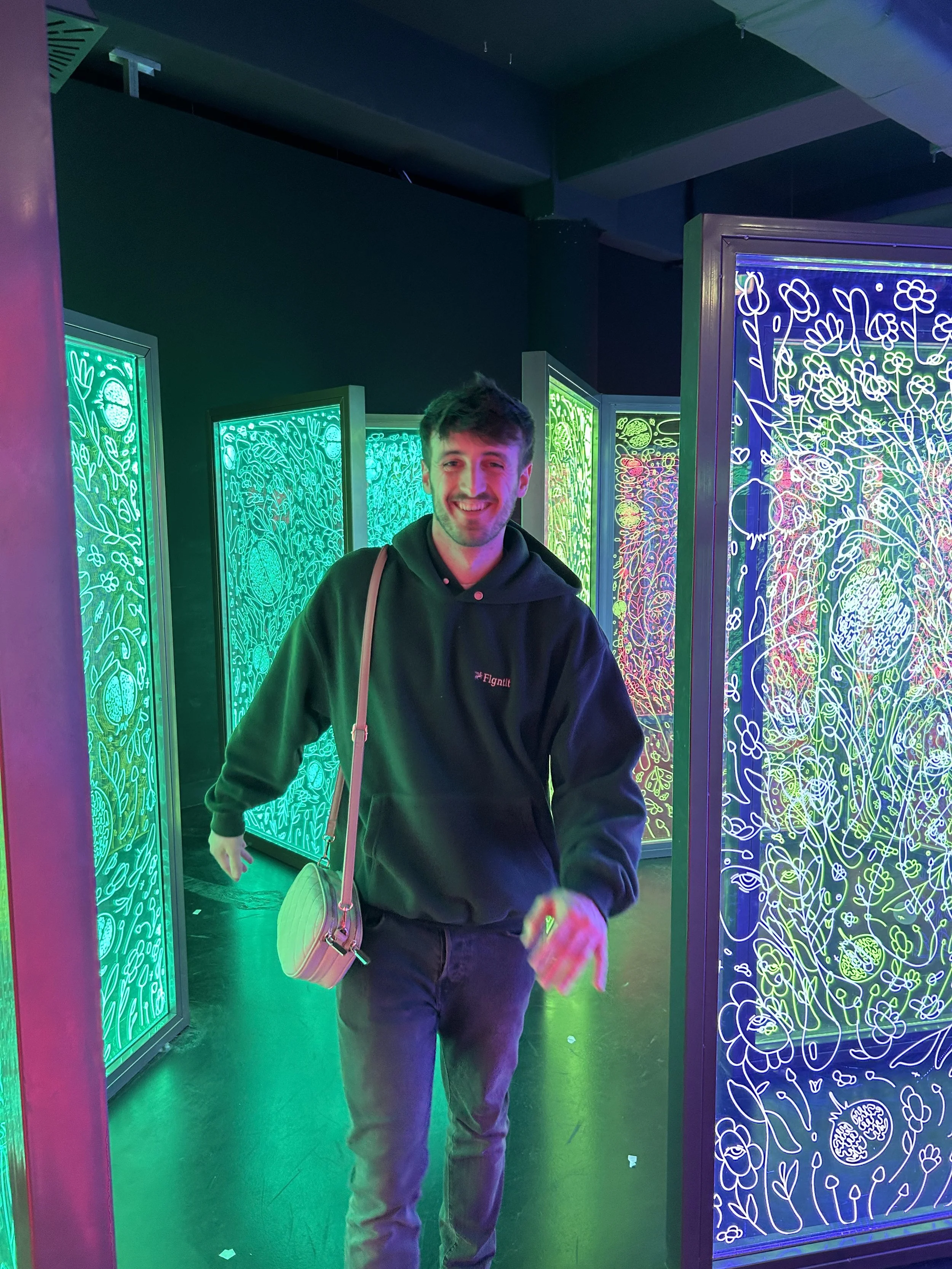 Junge Mann in dunklem Kapuzenpullover mit Tasche chequebookm Muster lacht in einem Raum mit leuchtenden, bunten Neon-Grafiken auf den Wänden.