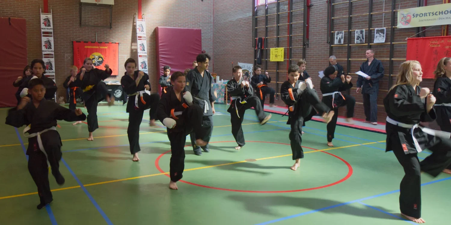 Kinderen oefenen karate in een gymzaal met instructeur, gekleed in zwarte karatepakken met gekleurde banden.