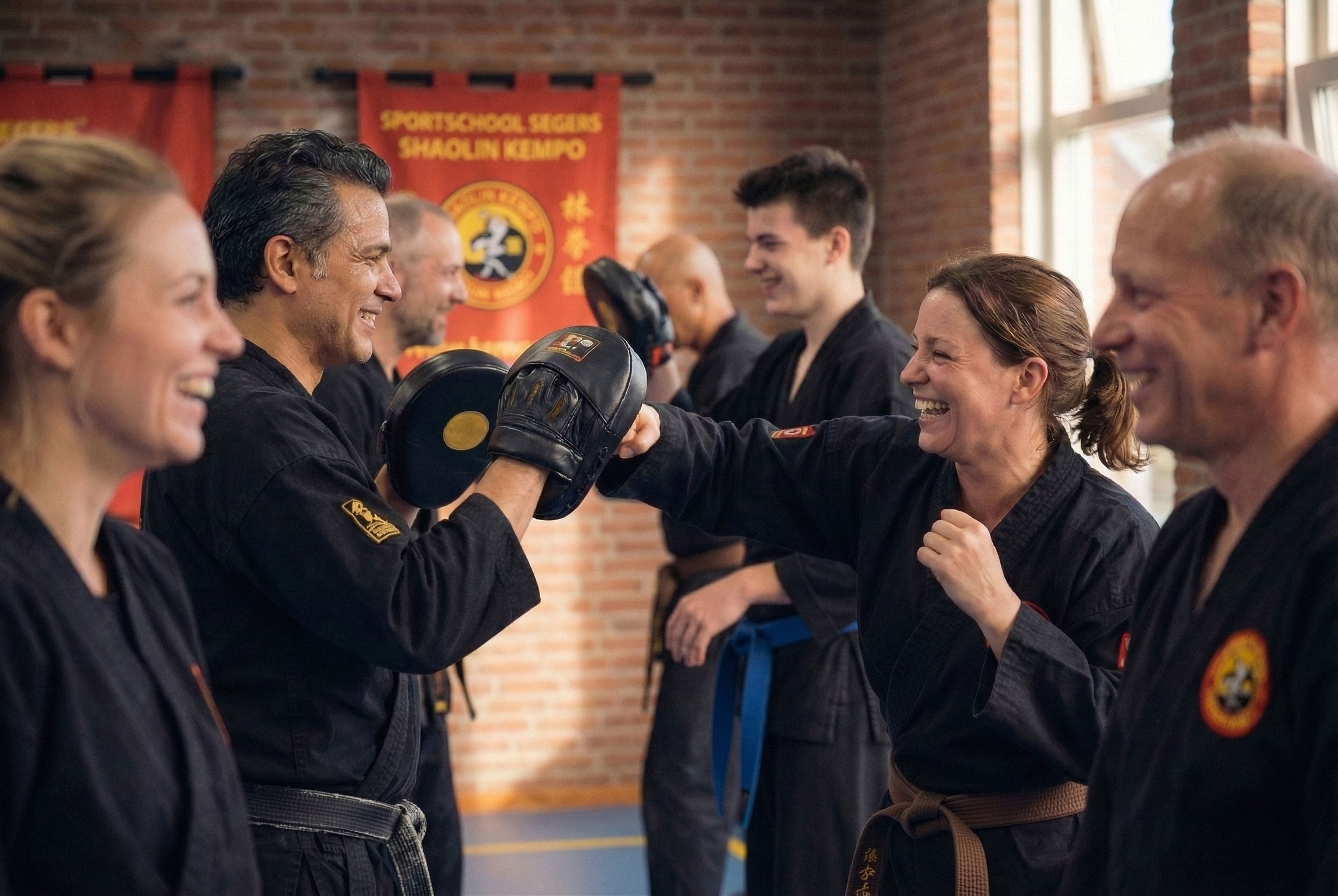 Mensen in een karate training, glimlachend en elkaar high-fivend, in een dojo met een banner op de muur.