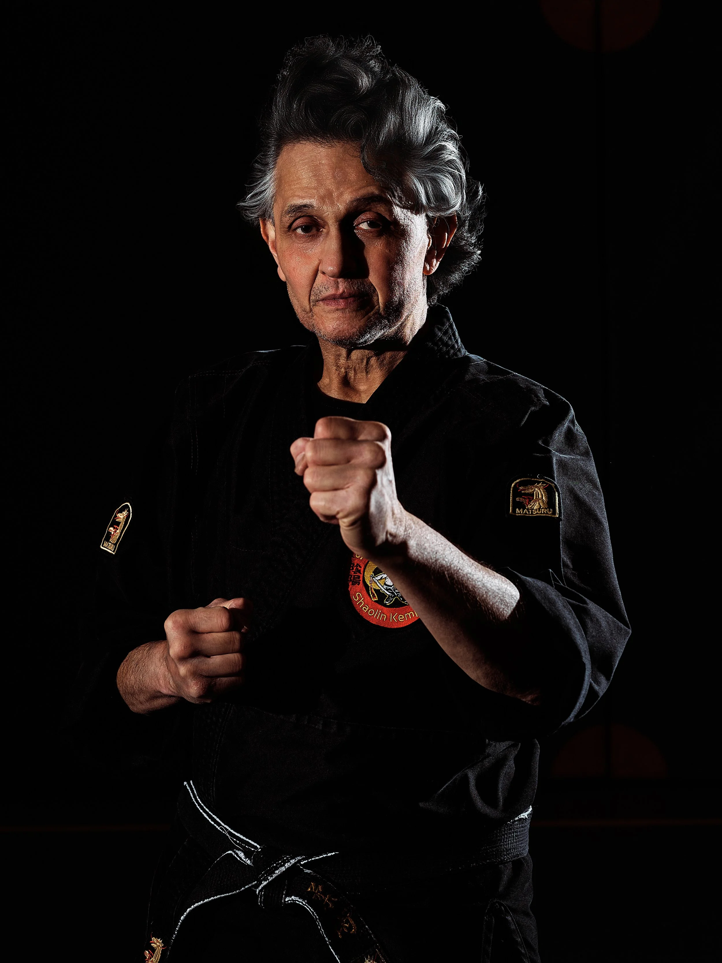 Een oudere man in een zwart karatepak met patches, poseert in een bokspositie.