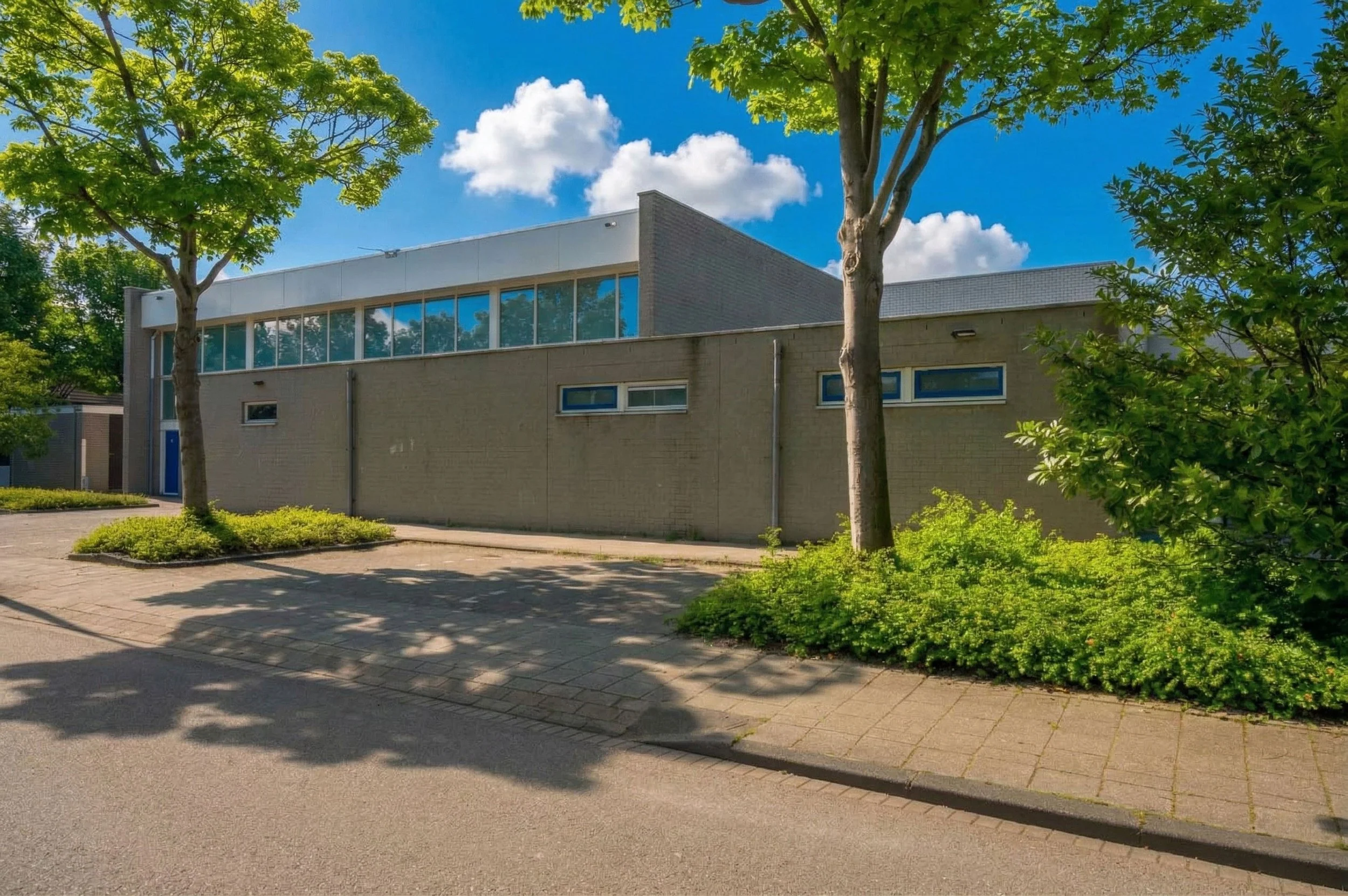 een modern gebouw met grote ramen en een lichtgele stenen muur, omgeven door groene bomen en struiken, onder een blauwe lucht met witte wolken.