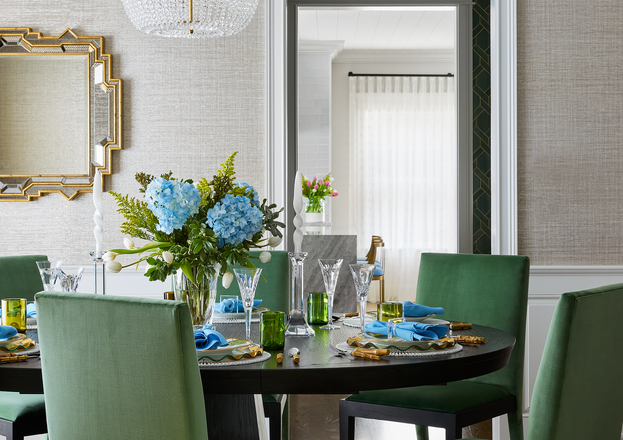 A dining room table decorated for a meal with a floral centerpiece featuring blue hydrangeas and white tulips, surrounded by green upholstered chairs, with place settings including blue napkins, glassware, and gold utensils, and a large mirror on the wall with a gold geometric frame.