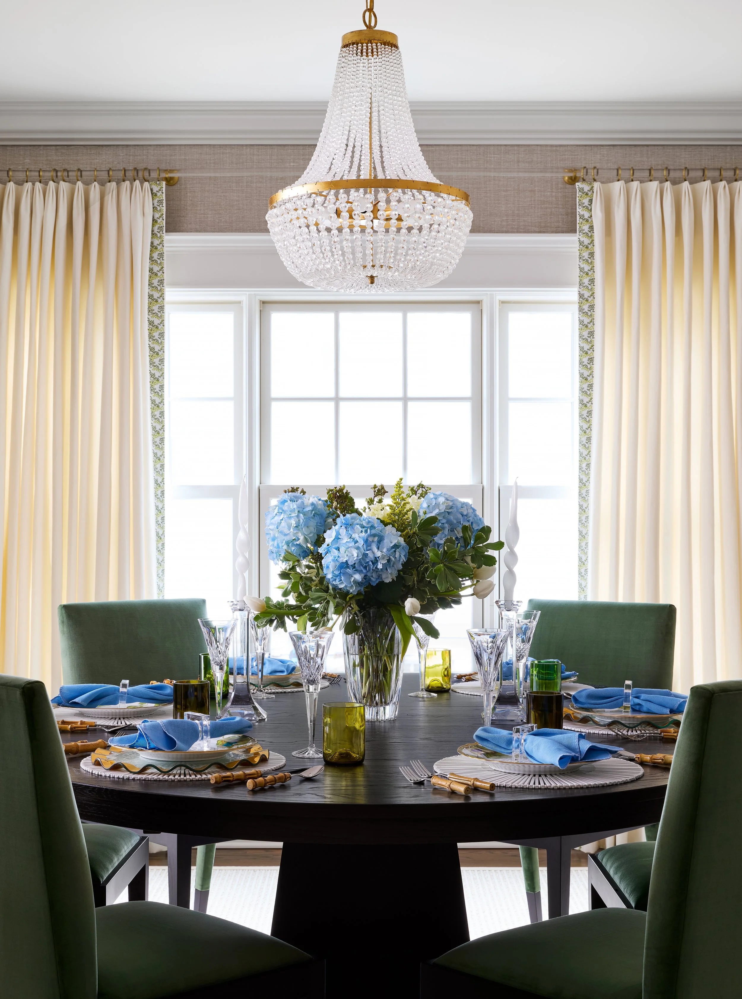 A dining room with a round black table set for six, featuring blue napkins, clear glassware, and a floral centerpiece of blue hydrangeas, with green velvet chairs, a large window, white curtains, and a crystal chandelier overhead.